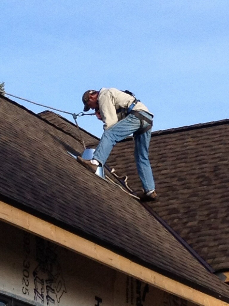 A person wearing a safety harness carefully works on a shingled roof, engaged in roofing tasks. They are bent over, handling tools or materials, securely fastened by ropes. The clear sky provides a perfect backdrop for their storm roof repair efforts.