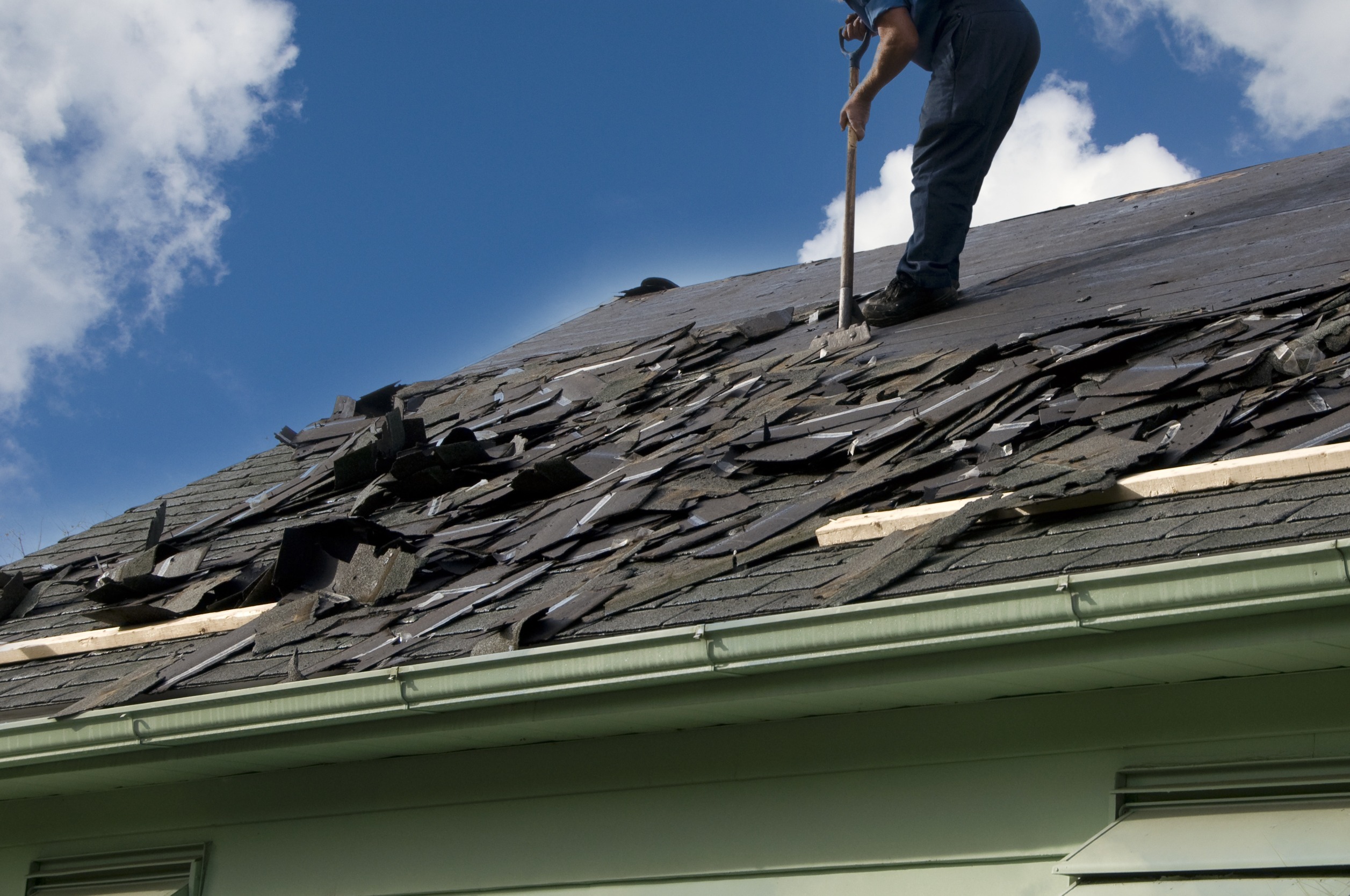 A person is engaged in storm roof repair, using a tool to remove old shingles. The sky is clear with a few clouds, creating a sunny setting for the work. The roof is partially covered with broken shingles and debris.