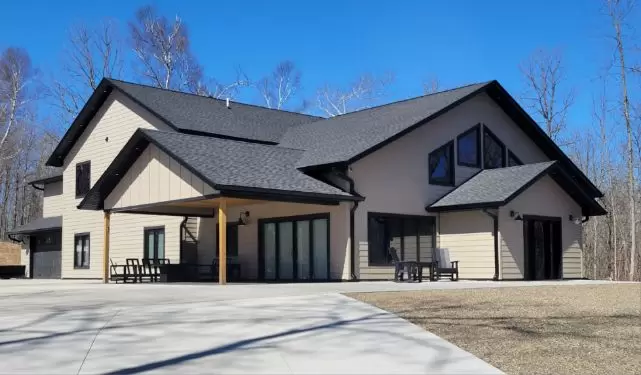 A modern, two-story house with beige siding and a dark gray roof, featuring large windows and a spacious patio with outdoor furniture. The home is surrounded by leafless trees under a clear blue sky.