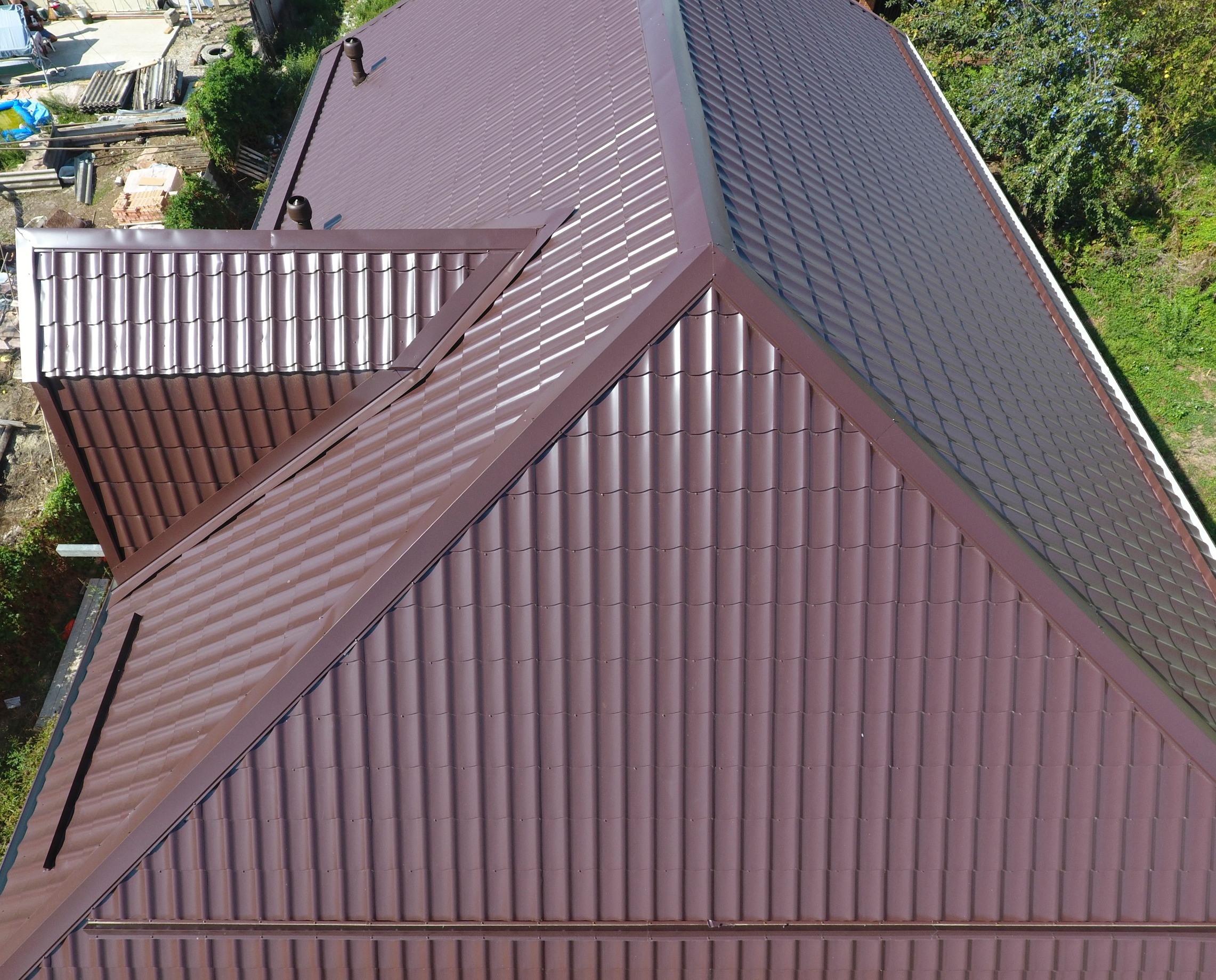 Aerial view of a building with a new brown metal roof. The roof has multiple peaks and valleys, featuring a ribbed pattern. Surrounding the building are trees, construction materials, and a partially built structure.