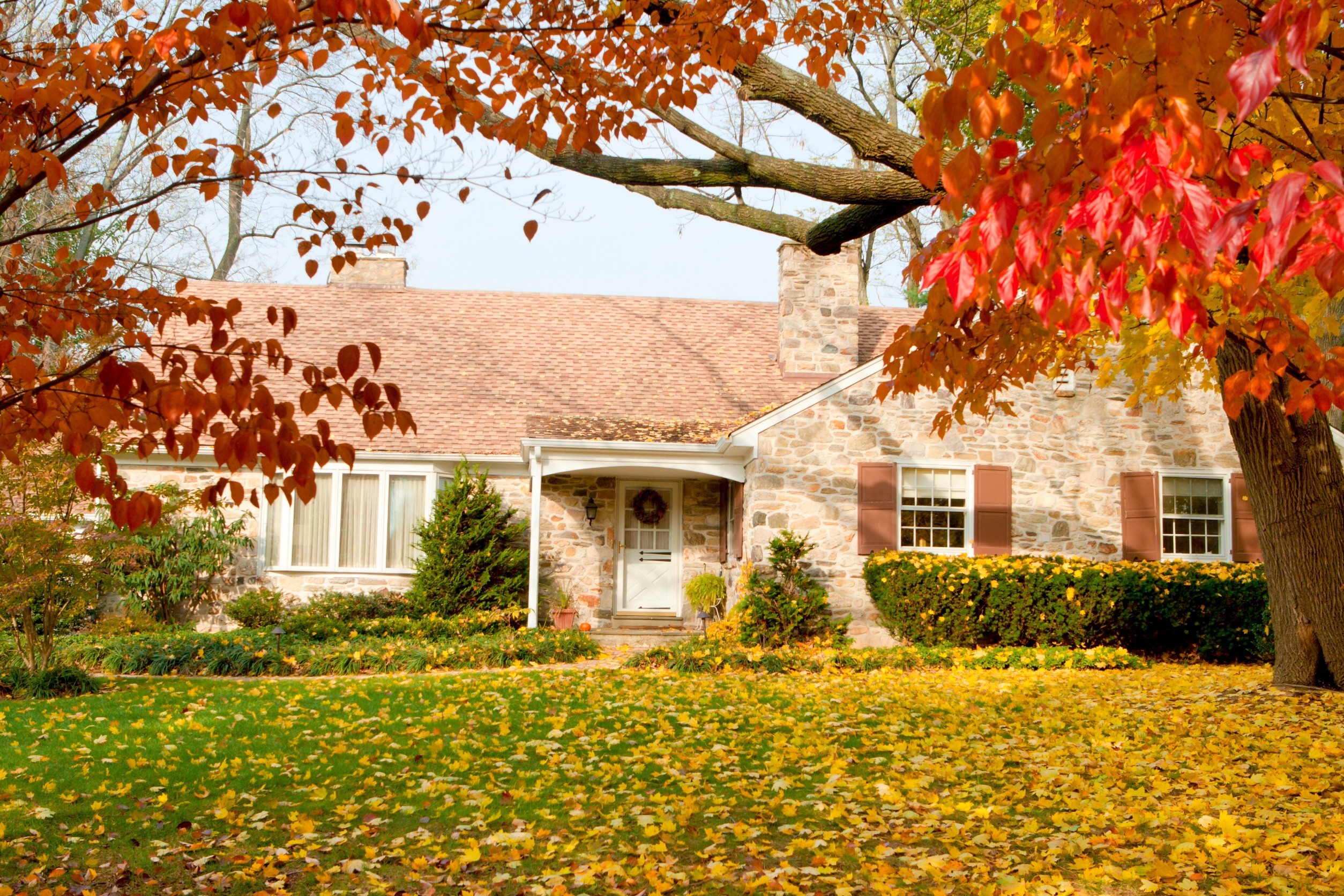 A cozy stone house with a chimney is surrounded by vibrant autumn foliage. Orange and red leaves frame the scene, with a lush green lawn covered in fallen leaves. A decorative wreath adorns the white front door.