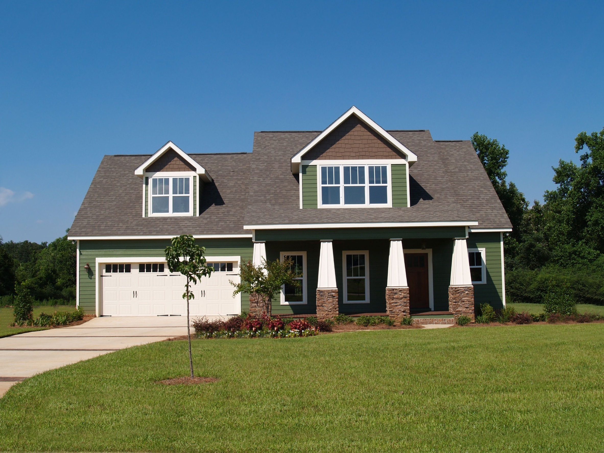 A modern green two-story house with white trim and a gray roof. It features a spacious lawn, a two-car garage, and a front porch with white columns, surrounded by lush greenery under a clear blue sky.