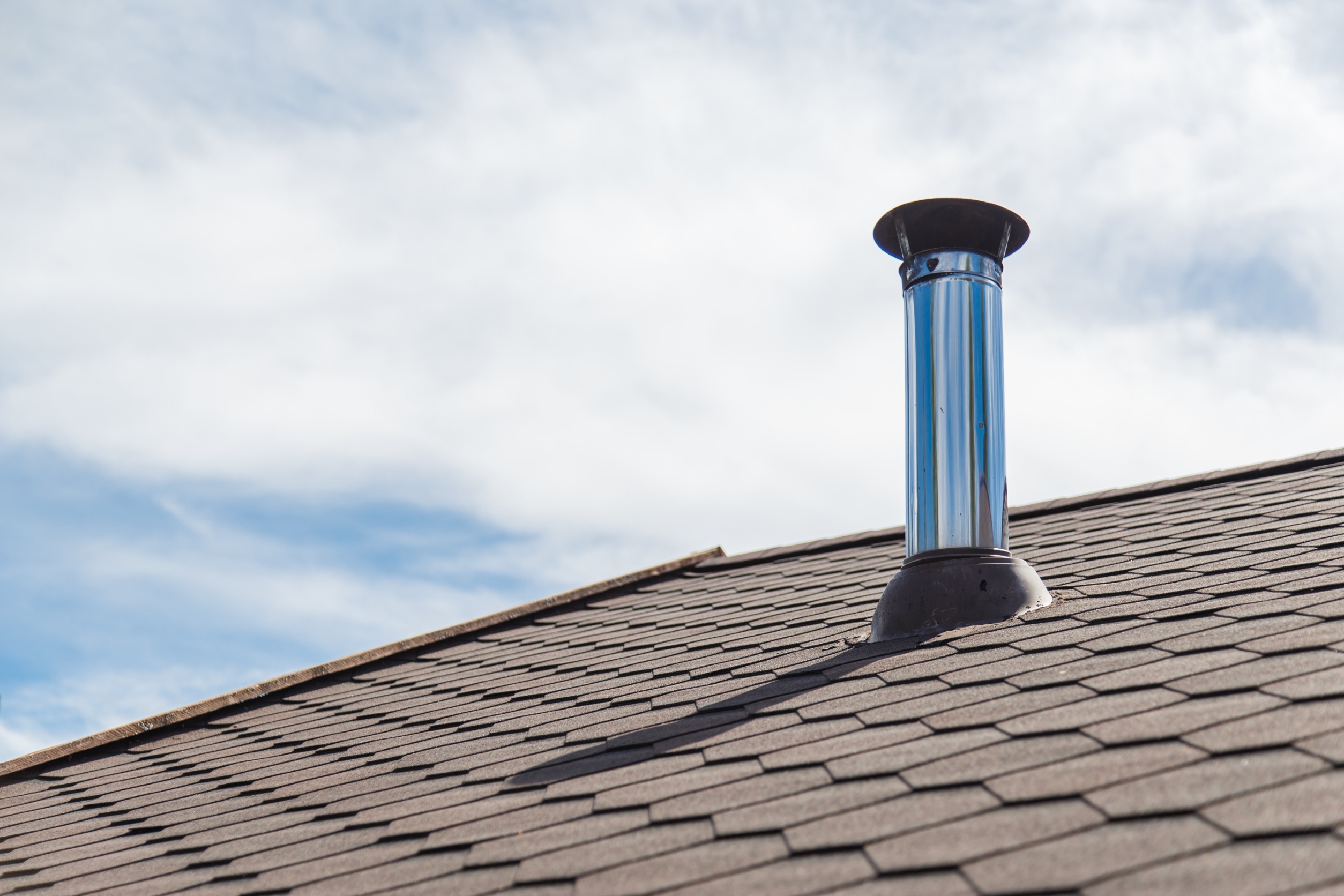 A rooftop with dark shingles and a shiny metal chimney pipe protruding from it. The sky in the background is partly cloudy and blue.