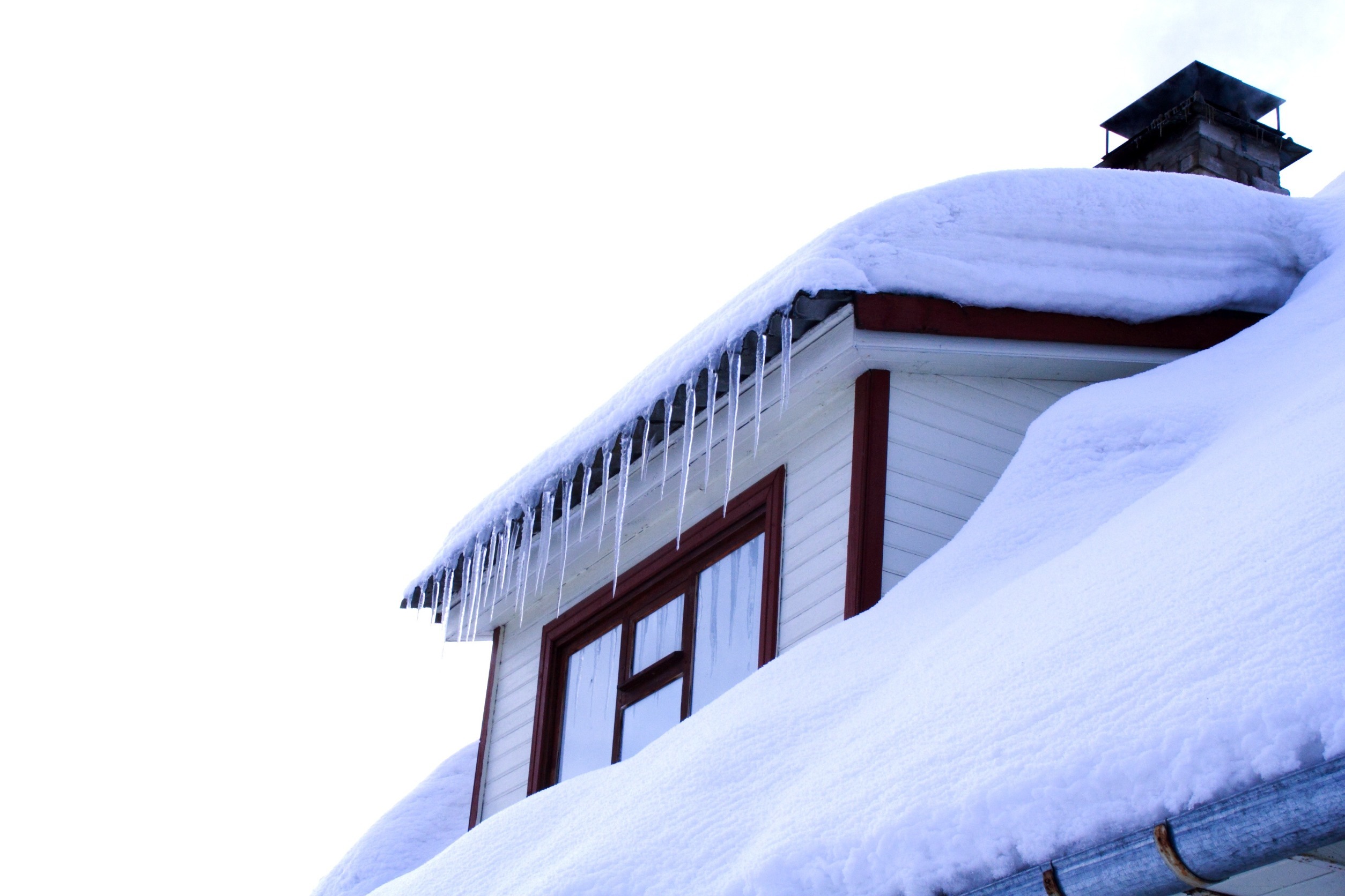 A snow-covered roof with a chimney and icicles hanging from the edge. The wooden house has a red-framed window, and the sky is bright and clear.