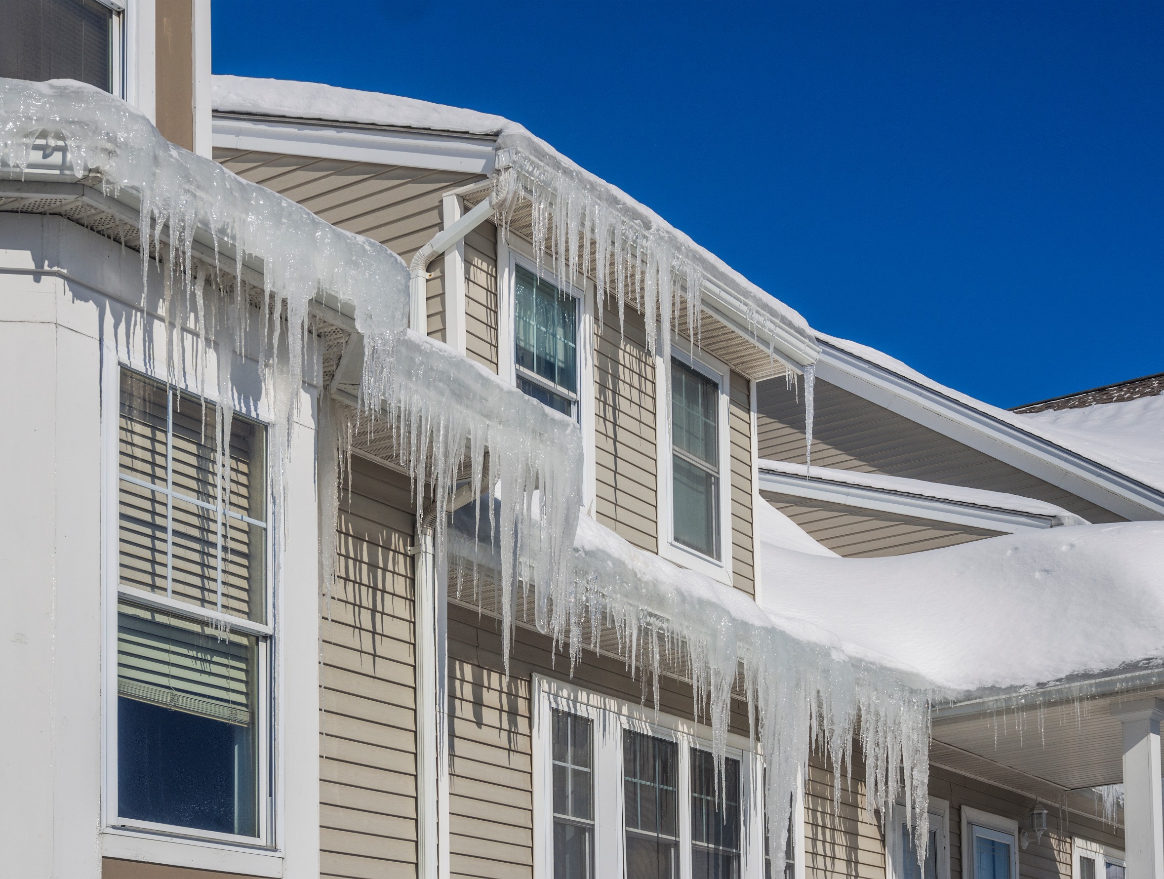 A house with light beige siding is adorned with long icicles hanging from the roof. The roof is covered in thick snow, and the sky is clear and bright blue. Sunlight reflects off the icicles, creating a shimmering effect.