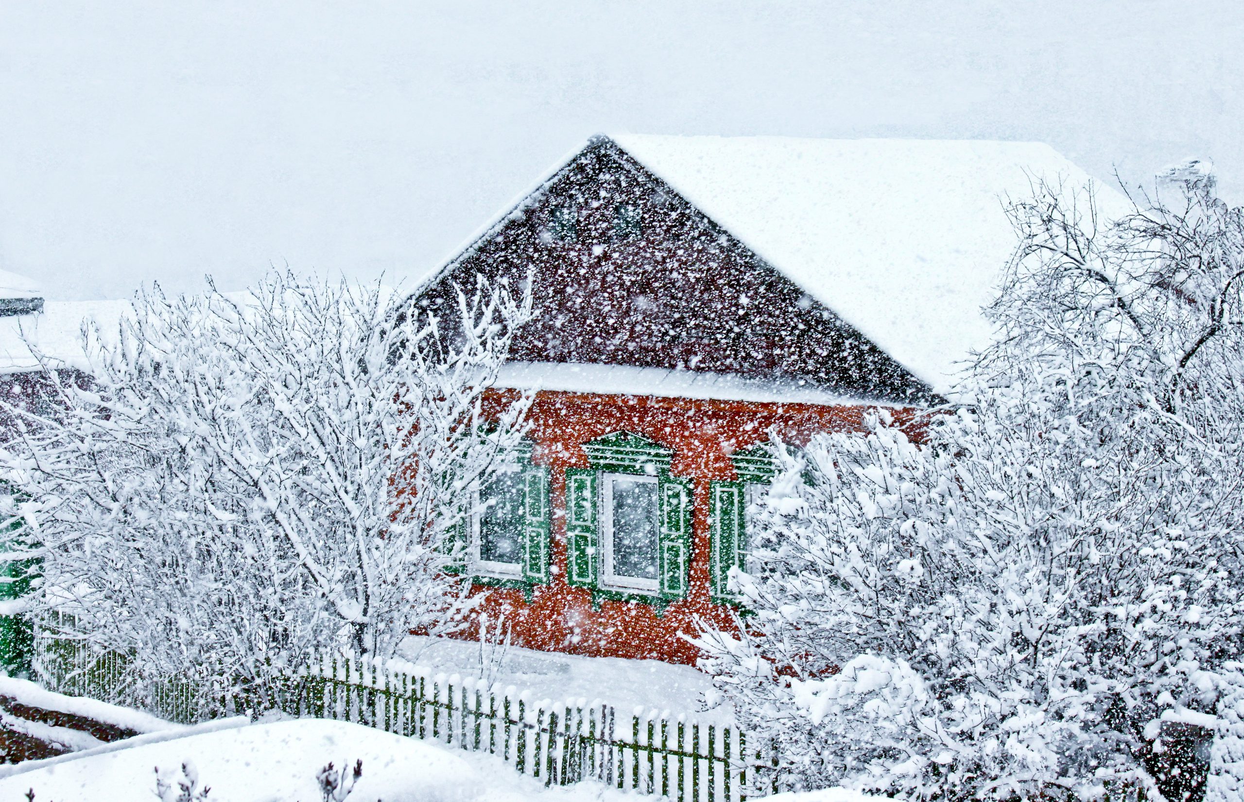 A quaint red house with green shutters and a snow-covered roof stands amidst a heavy snowfall. Leafless trees and a wooden fence surround the house, all blanketed in snow, creating a serene winter scene.