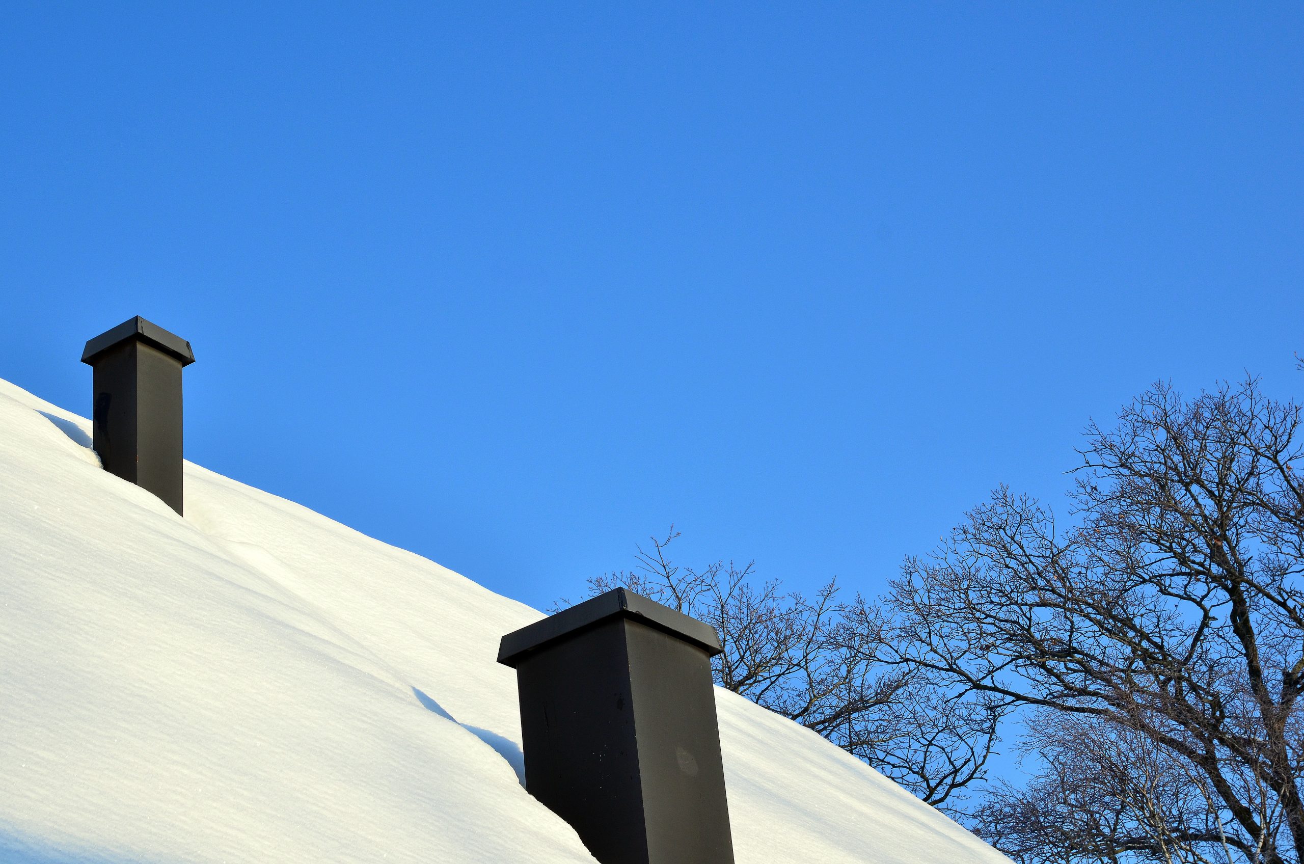 A snowy rooftop with two black chimneys against a clear blue sky. Bare tree branches are visible in the background.