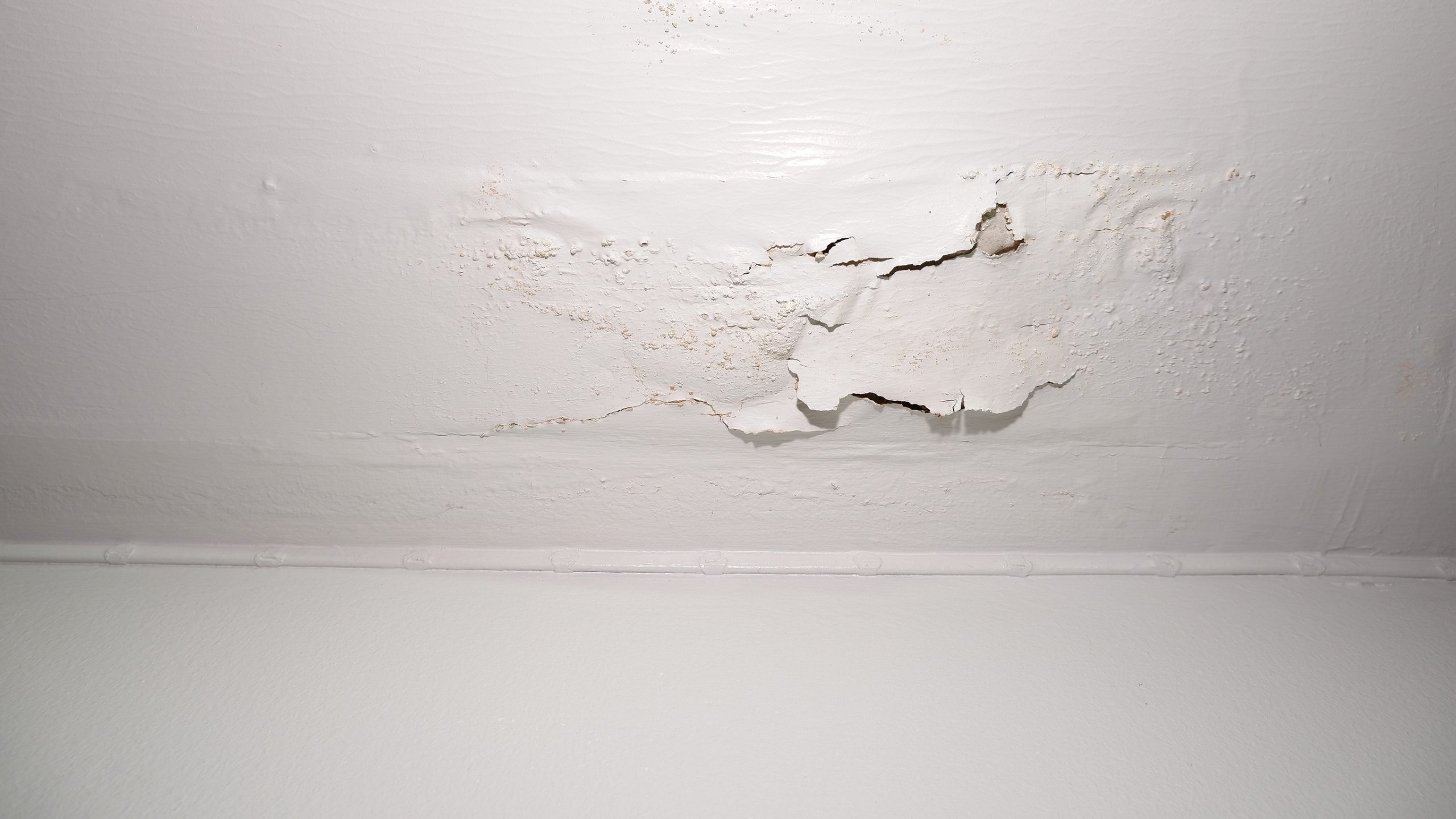 A white ceiling with peeling, cracked paint and visible water damage above a smooth wall.