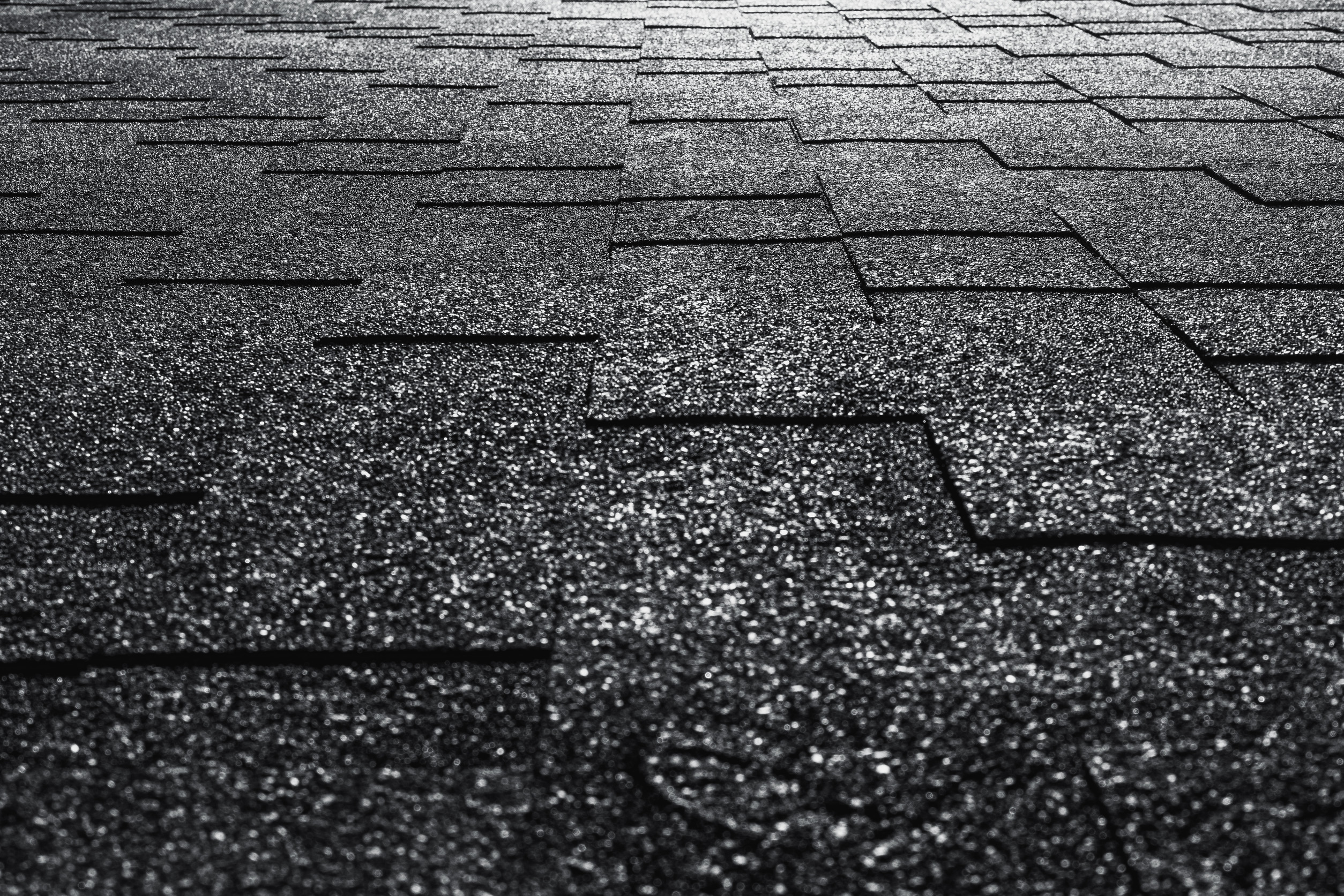 Close-up view of dark asphalt roof shingles with a textured, rough surface. The shingles are arranged in a staggered pattern, and sunlight creates reflective highlights across the surface.