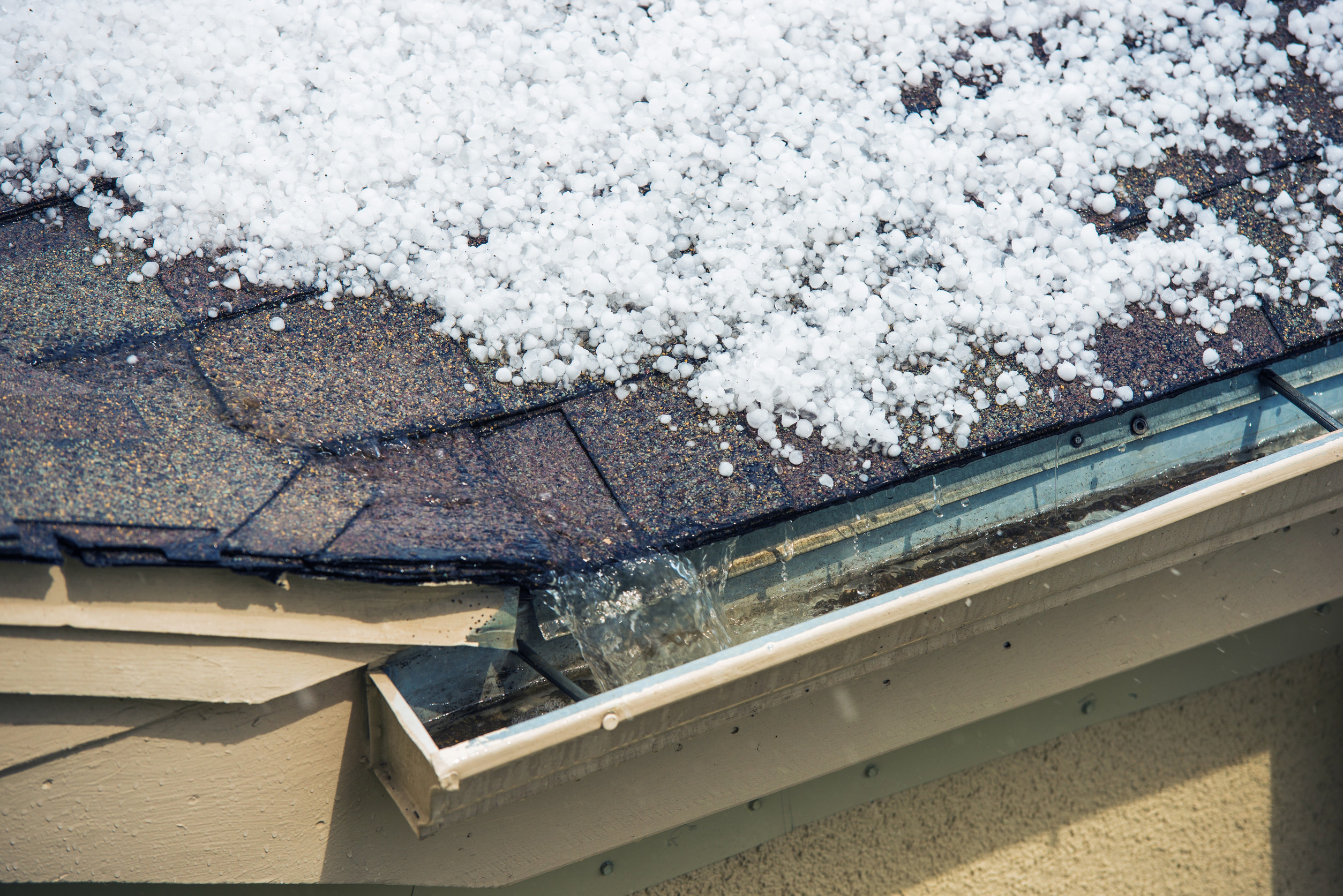 Hailstones covering the roof and filling the gutter of a house, with water visibly flowing from the gutter, indicating a recent hailstorm.