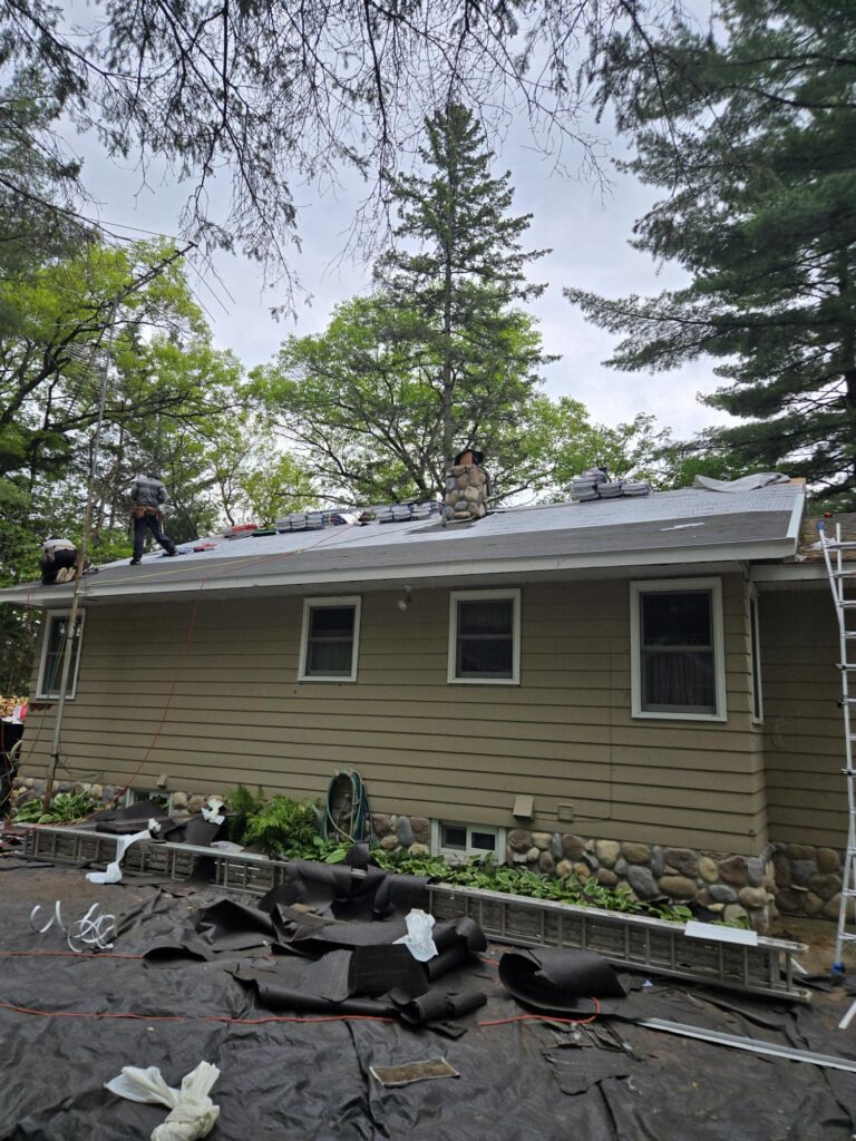 Several workers repair or install a roof on a tan house with white trim surrounded by trees. Ladders are set up against the house, and roofing materials are scattered on the ground below.