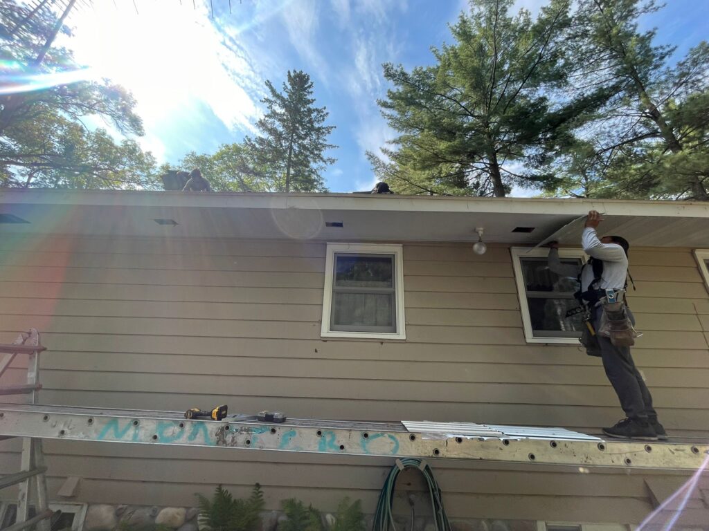 A worker on a ladder repairs or installs siding near the roof of a beige house, while another worker is visible on the roof. Trees and sunlight with lens flare are in the background. Tools are scattered nearby.