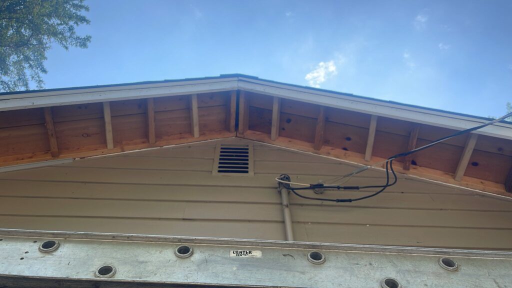 View looking up at the gable end of a house with exposed rafters, beige siding, a small vent, visible cables, and a metal ladder positioned at the base. The sky above is clear with a few clouds.