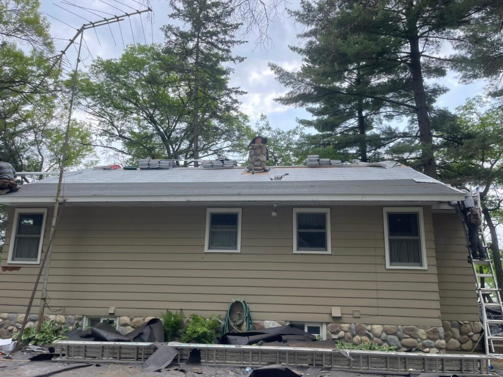 A tan house with a stone chimney and a partially completed roof renovation. Roofing materials and workers are visible on the roof. Trees and cloudy sky are in the background.