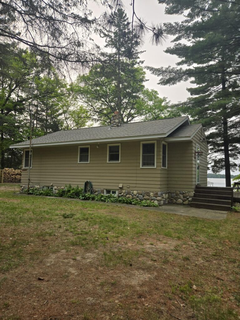 A small beige house with a stone foundation sits among tall pine trees. There are three windows on the side, a hose reel, and a stack of firewood in the background. A pathway leads to steps at the back entrance.
