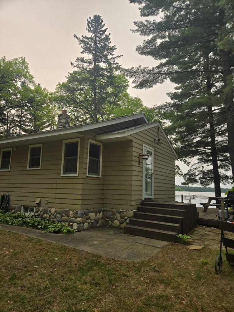 A small beige cabin with a stone foundation sits near tall trees, wooden steps leading to the entrance, and a lake visible in the background through the trees.