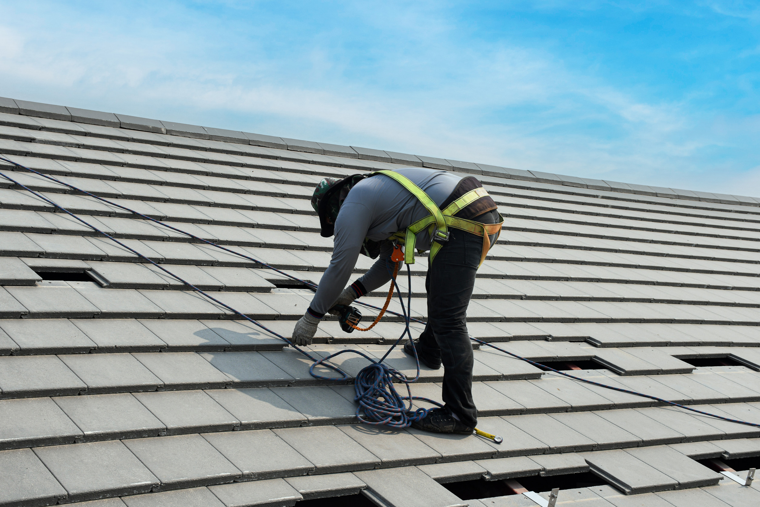 A construction worker wearing safety gear and a harness is installing tiles on a sloped roof under a clear blue sky, using tools and ropes for safety and support.