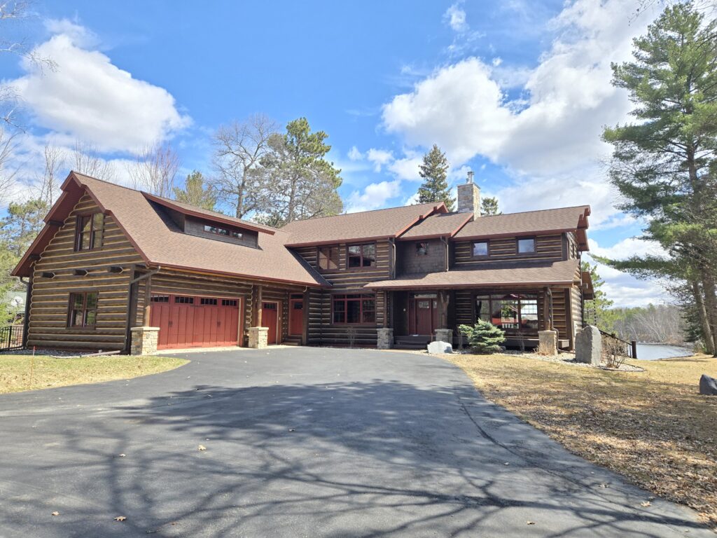 A large log cabin-style house with a red roof and garage doors, surrounded by trees and a paved driveway, set under a blue sky with scattered clouds. A lake is visible in the background to the right.