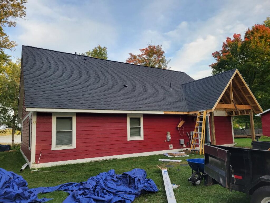 A red house with white trim is under renovation, featuring a newly shingled black roof. Ladders, tarps, and construction materials are scattered on the grass, and trees with autumn leaves are in the background.