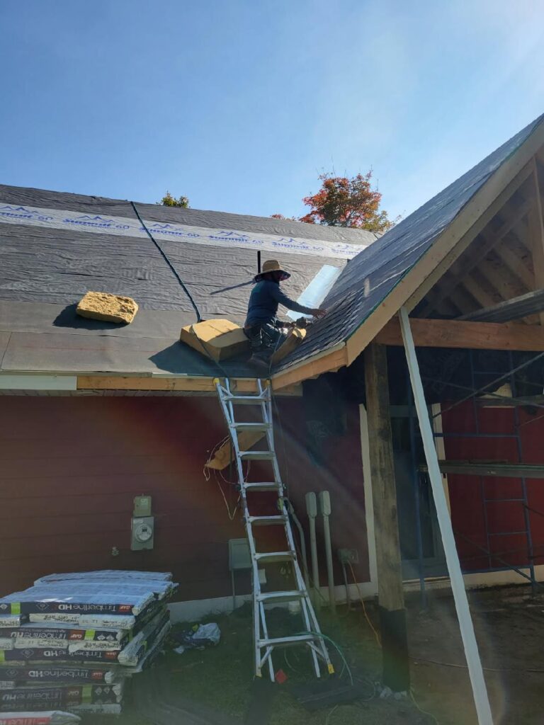 A worker on a ladder installs material on a sloped house roof under bright sunlight. Roofing supplies are on the ground, and the roof is partially covered with a black underlayment. Scaffolding stands nearby.