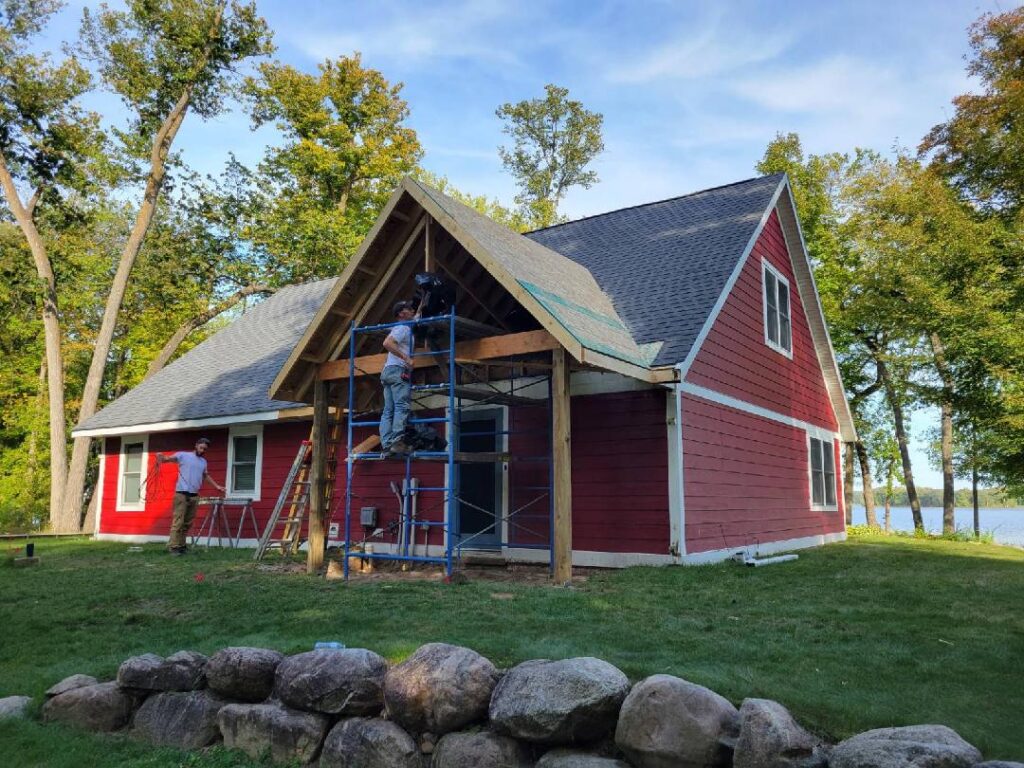 Three workers are building a wooden porch on a red house with white trim. Scaffolding is set up under the porch roof, and tall trees and a lake are visible in the background. Large rocks border the foreground.