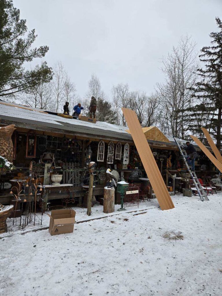 Several people work on the snow-covered roof of a rustic building surrounded by various antiques and metal decor items, with tall orange roofing panels propped against the structure and trees in the background.