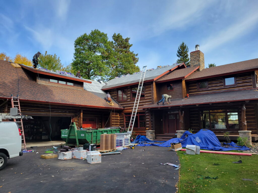 Workers are repairing the roof of a large log house. Ladders, tools, and materials are scattered on the driveway and lawn. A white van and green dumpster are parked nearby. Tarps cover parts of the ground and roofing area.