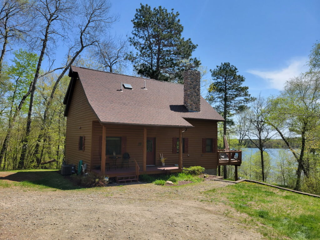 A wooden cabin with a stone chimney sits among trees near a lake, under a clear blue sky. There is a small deck on the side and a porch with chairs in front. The ground is partly grassy, partly gravel.
