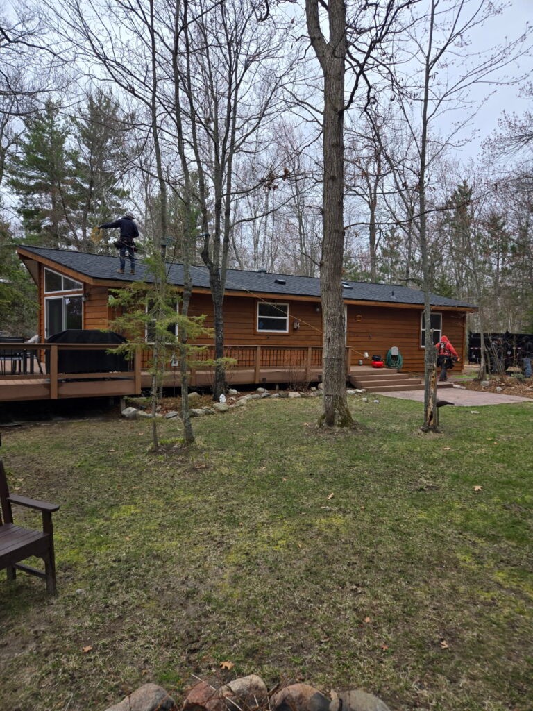 A wooden cabin surrounded by trees with a person on the roof and another person near the deck. The yard has green grass, a fire pit, and chairs, with forested woods in the background under a cloudy sky.