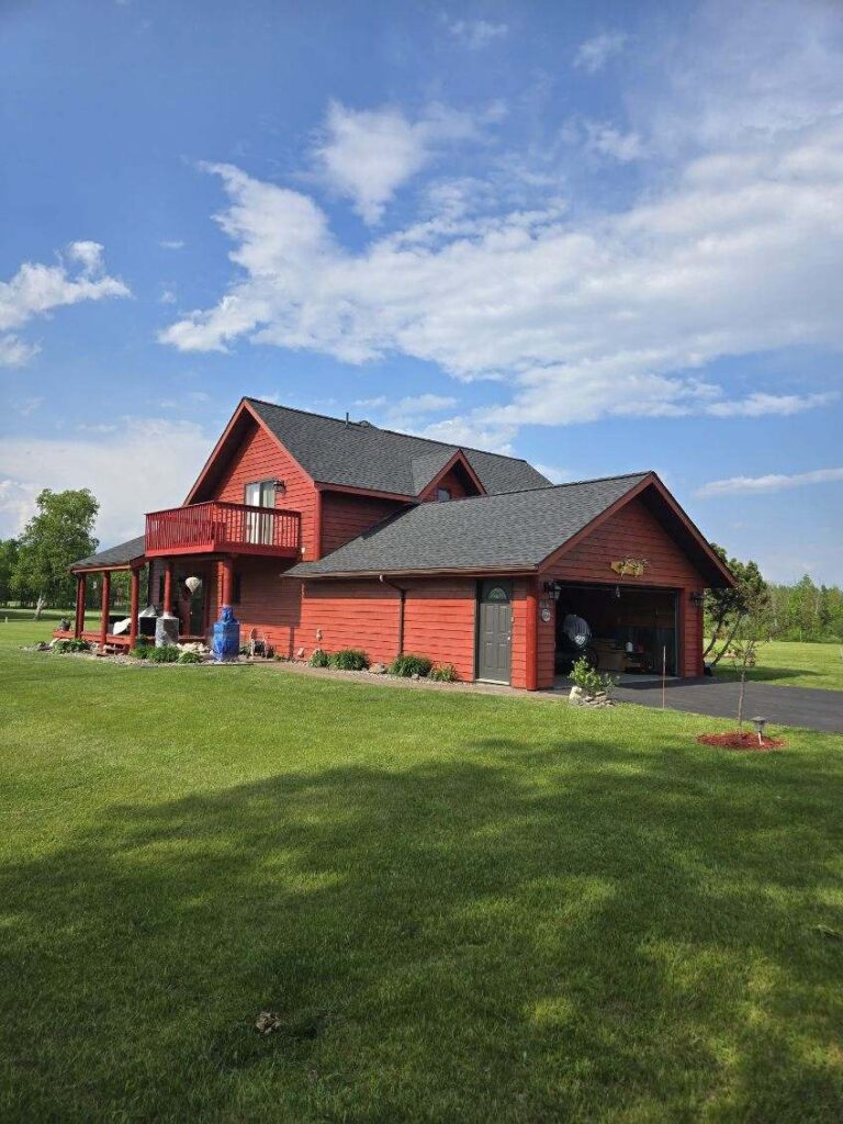 A red two-story house with a dark gray roof sits on a green lawn under a partly cloudy blue sky. There are flower beds, a small tree, and outdoor seating visible on the porch.