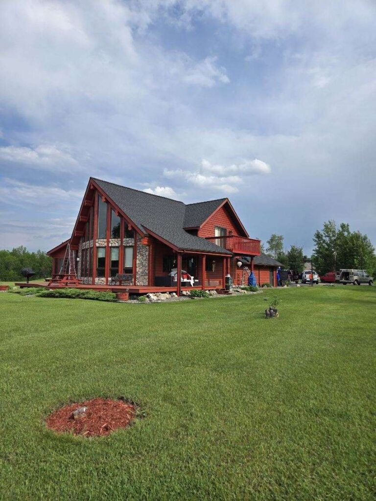 A red wooden cabin with large windows and a sloped roof sits on a well-maintained green lawn under a partly cloudy sky. Trees and parked vehicles are visible in the background.