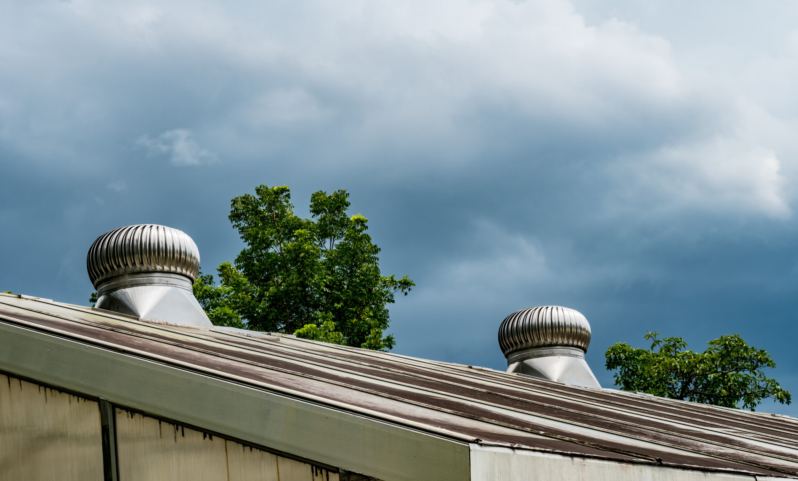 A slanted metal roof with two silver turbine vents is shown, with green trees and a cloudy sky in the background.