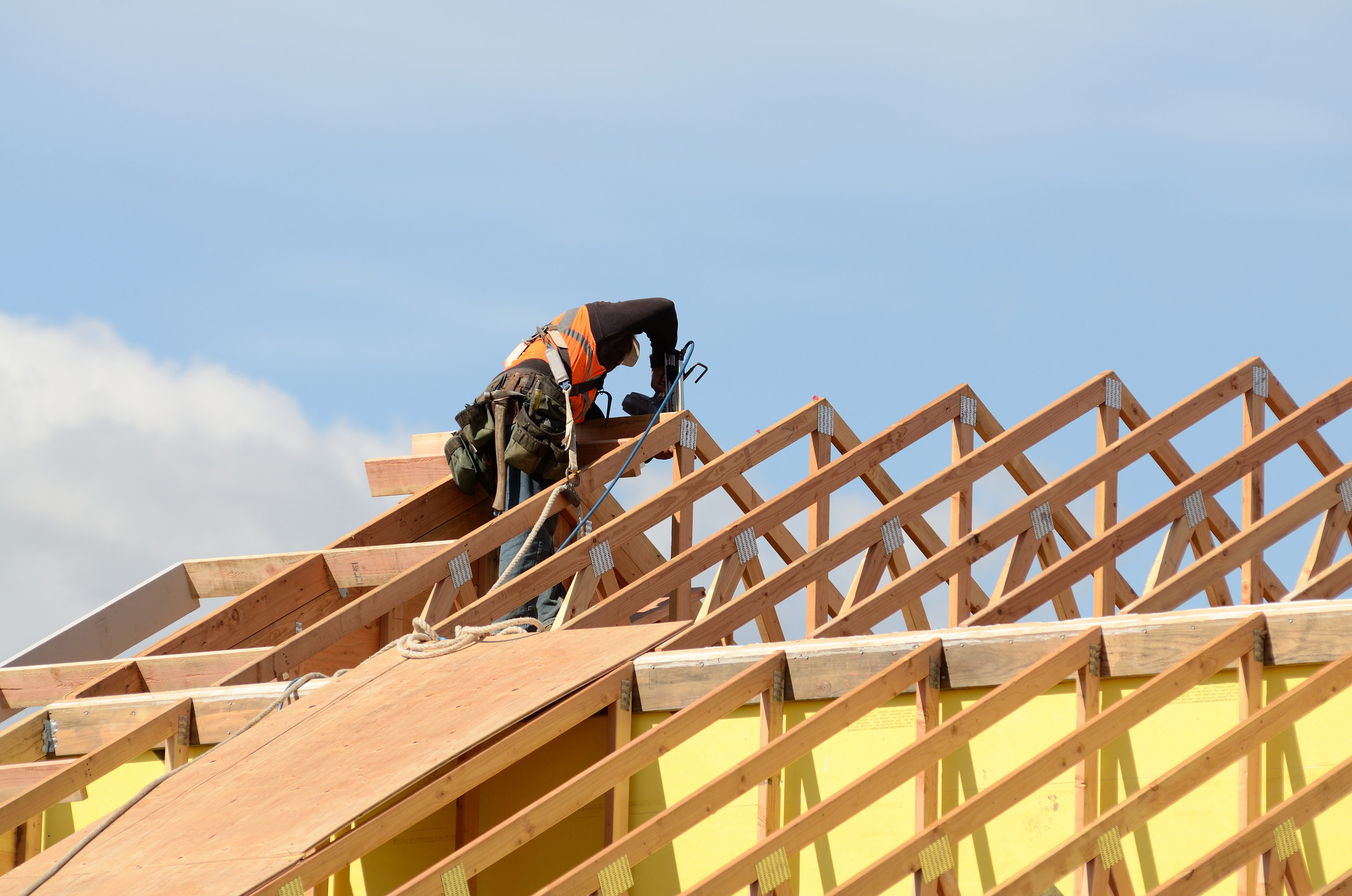 A construction worker wearing a safety harness and orange vest works on the wooden frame of a roof under clear blue sky.