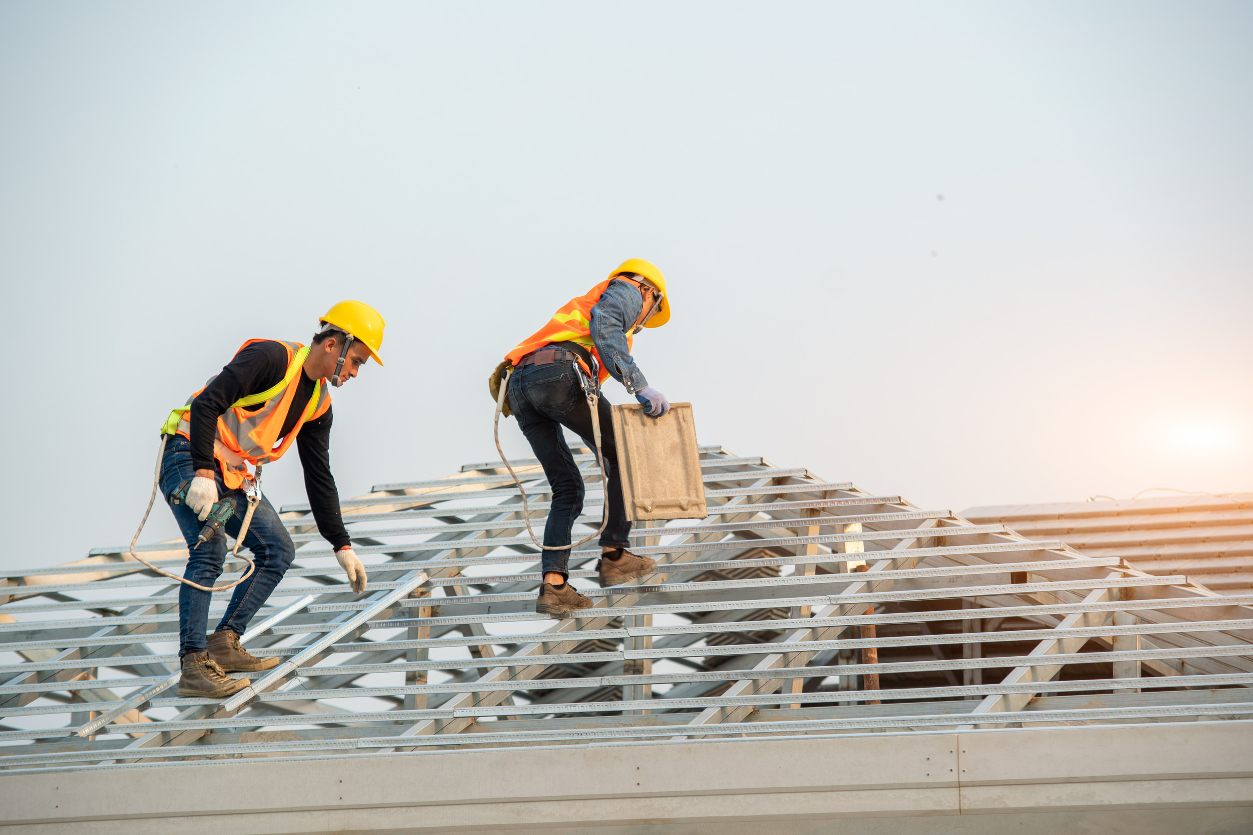 Two construction workers wearing safety vests and helmets work on a metal roof frame. One is crouching while the other is carrying a wooden board, both secured with safety harnesses against a clear sky background.