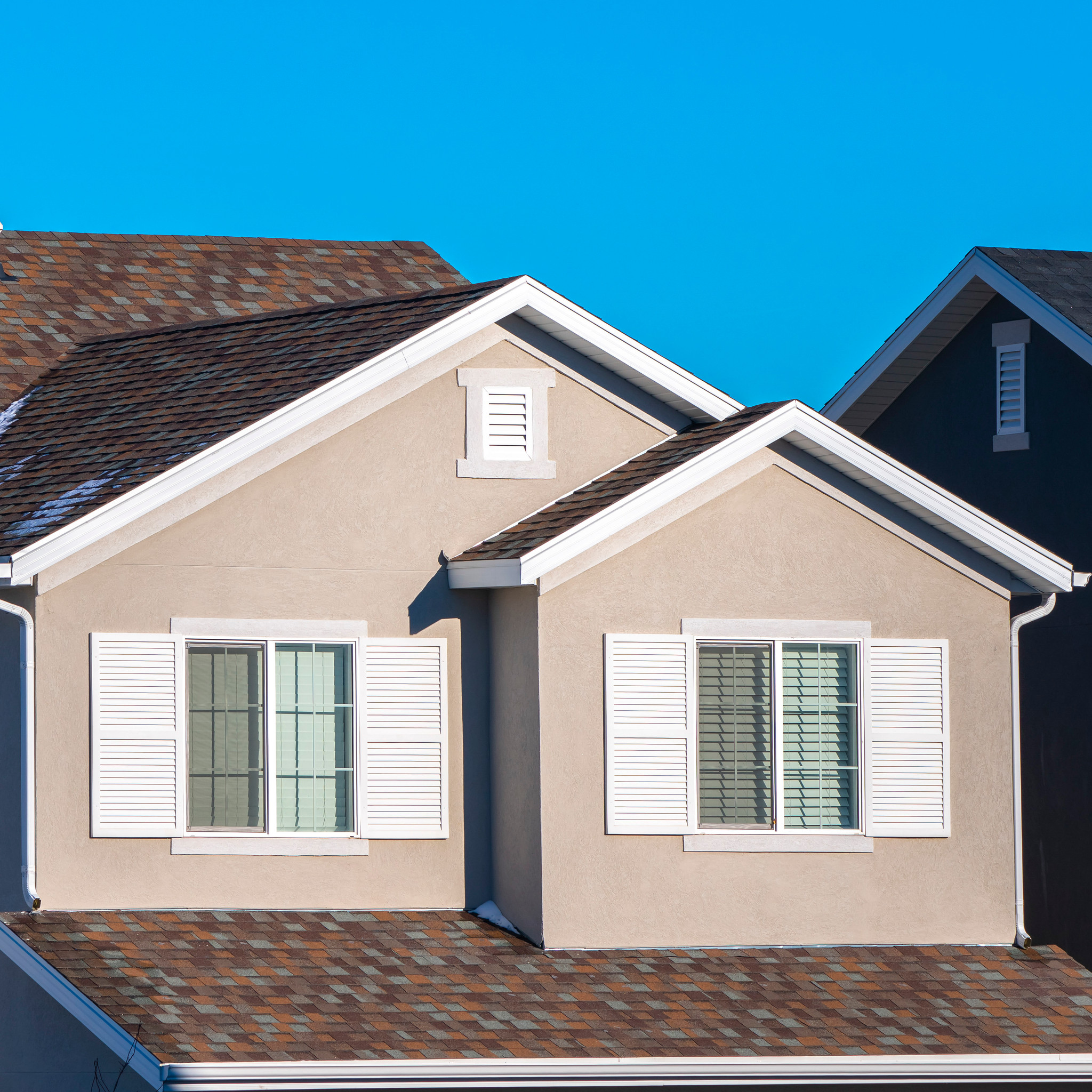 A beige house with white shutters and a brown shingle roof, featuring two large front-facing windows, under a clear blue sky.