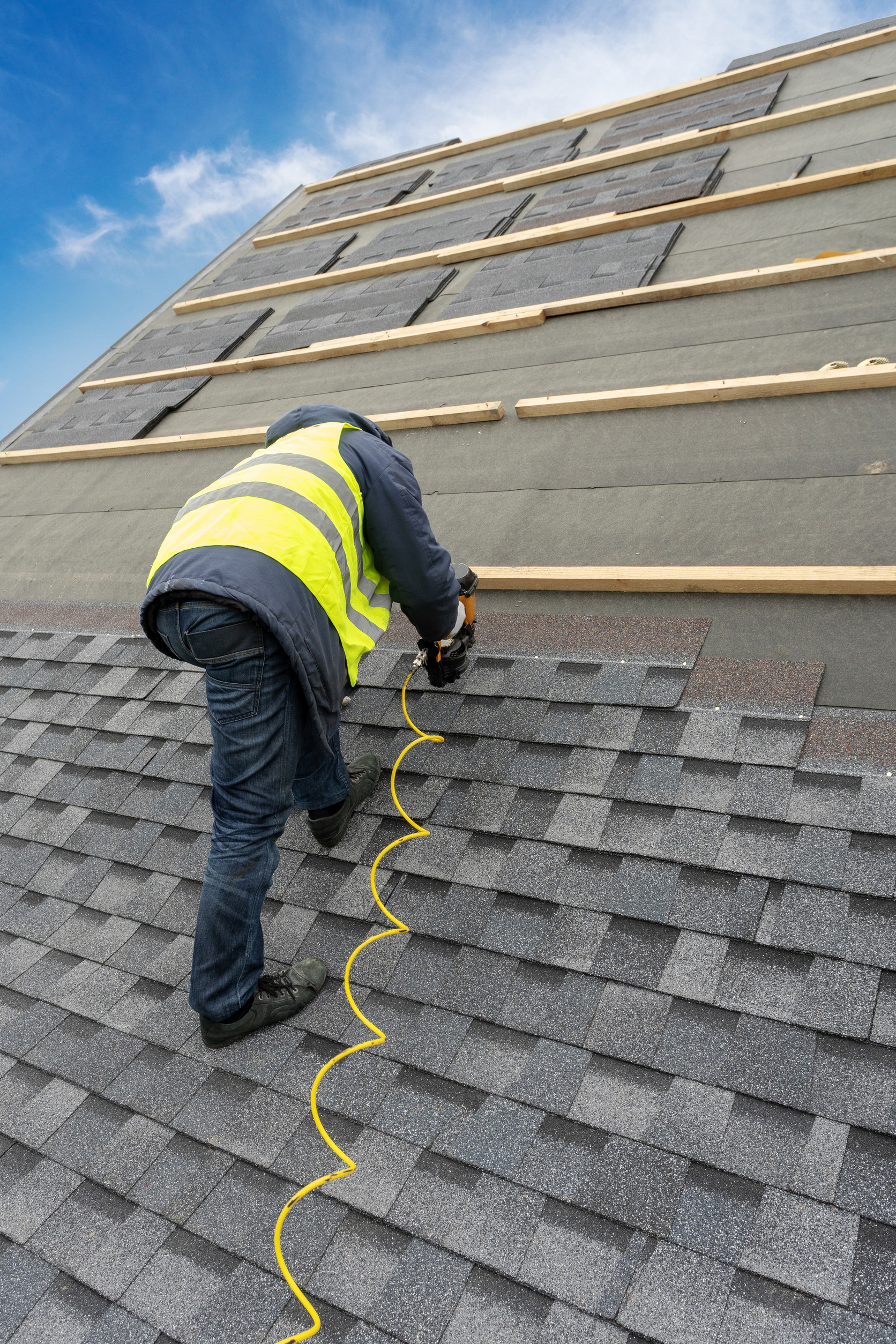 A worker in a high-visibility vest installs asphalt shingles on a sloped roof using a nail gun, with unfinished sections and wooden battens visible above. The sky is partly cloudy.