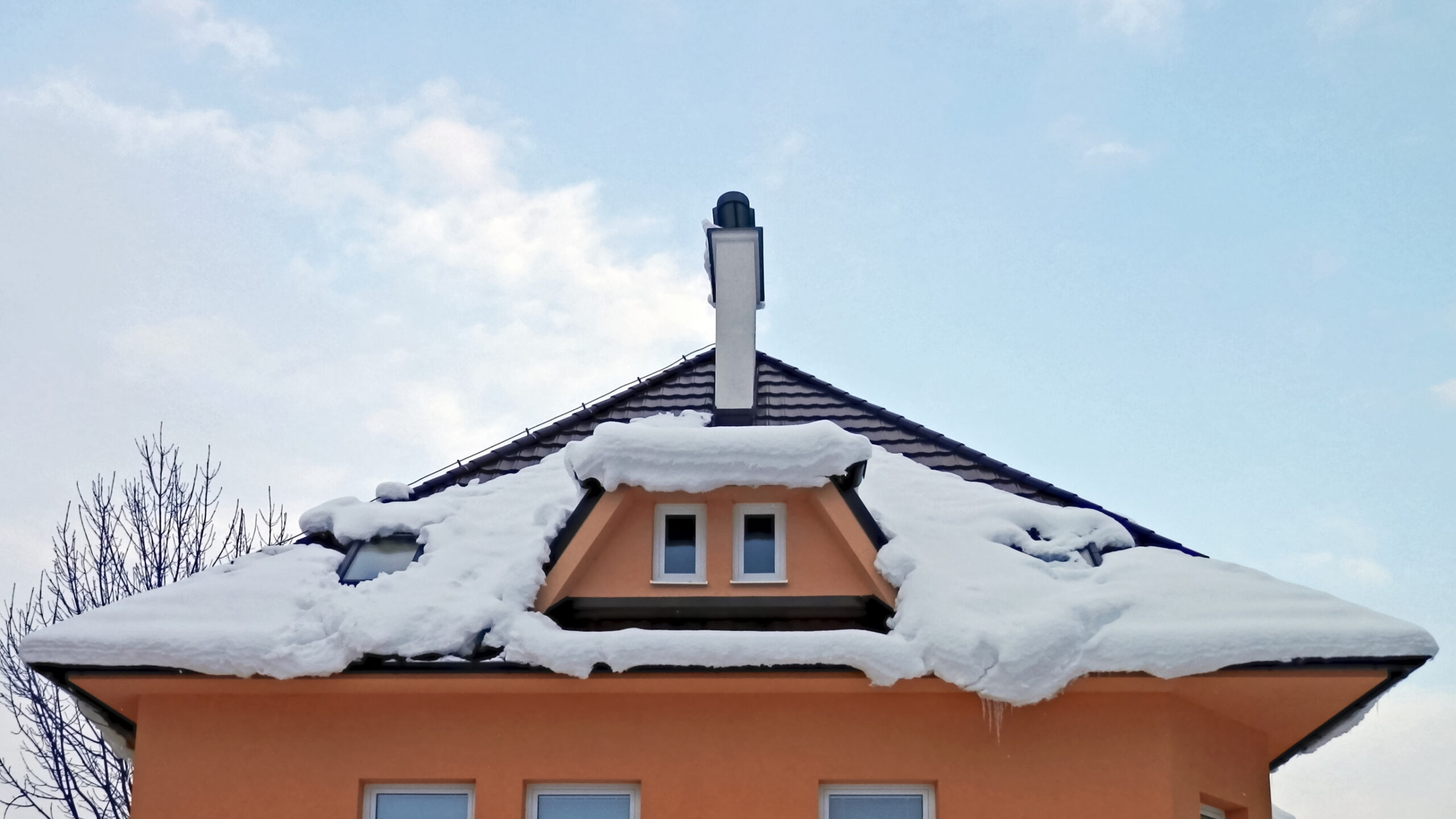 A house with an orange exterior and a snow-covered dark roof. Two small windows are set in the attic, and a white chimney rises above the roof against a partly cloudy sky.