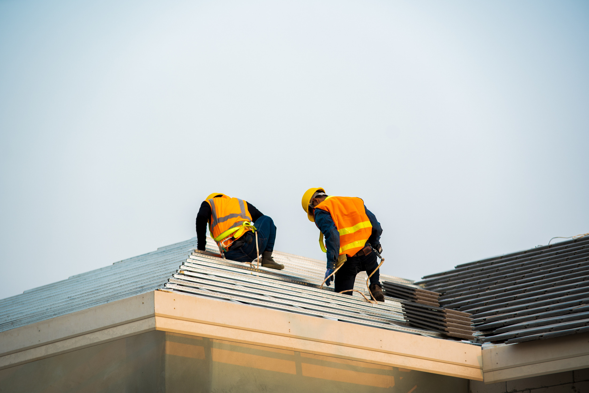 Two construction workers in orange safety vests and yellow helmets work on installing metal roofing on a building under clear sky.
