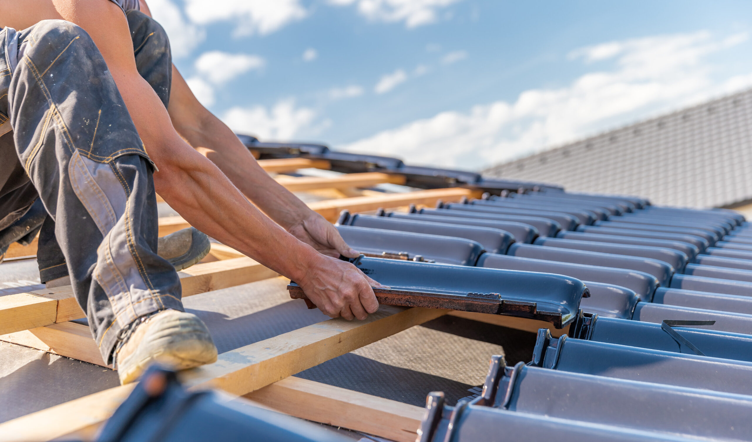 A construction worker installs dark roof tiles onto wooden beams under a bright, clear sky, assembling the roofing structure of a building.