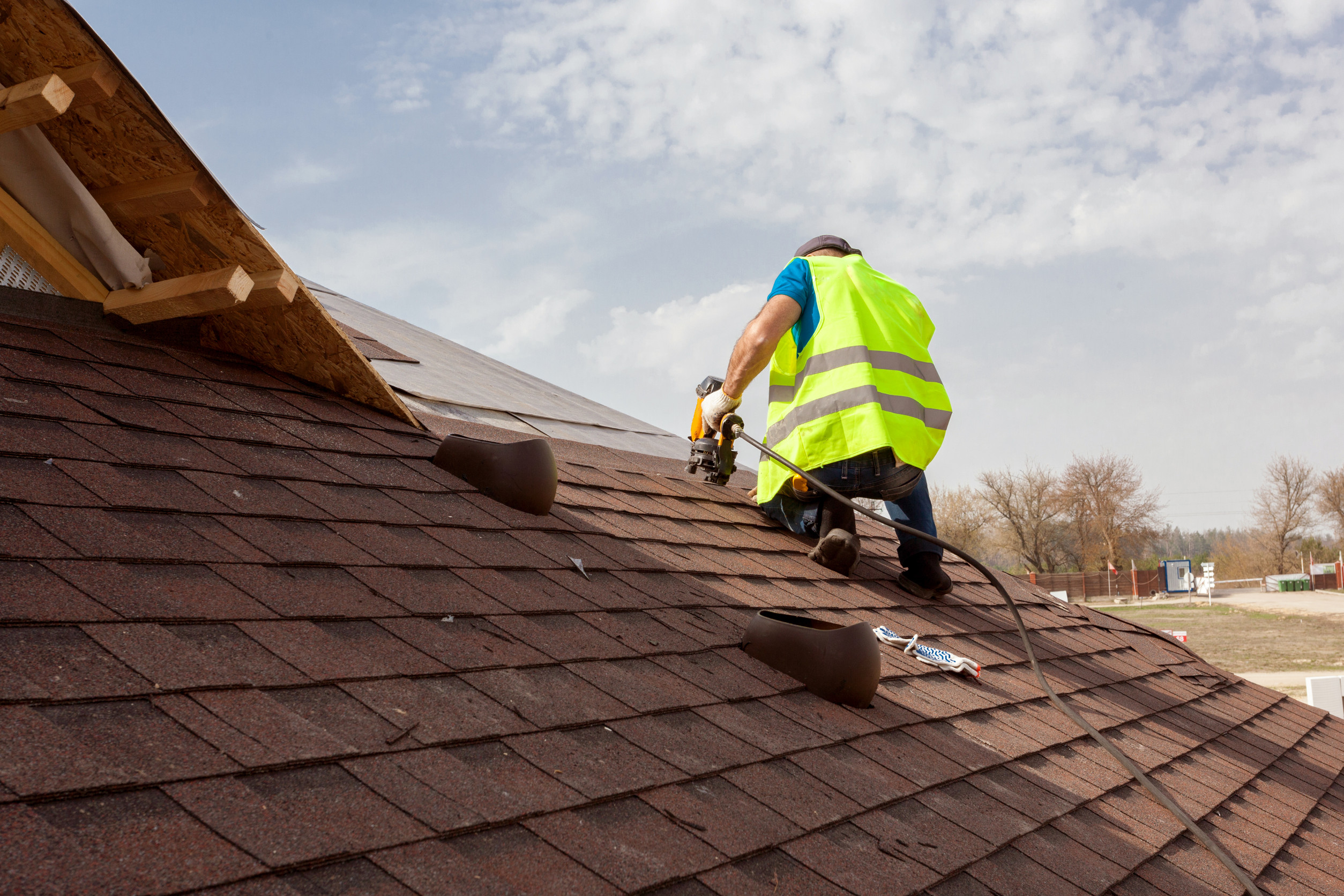 A worker in a neon safety vest installs shingles on a sloped roof using a power tool, with roofing materials and trees visible in the background under a partly cloudy sky.
