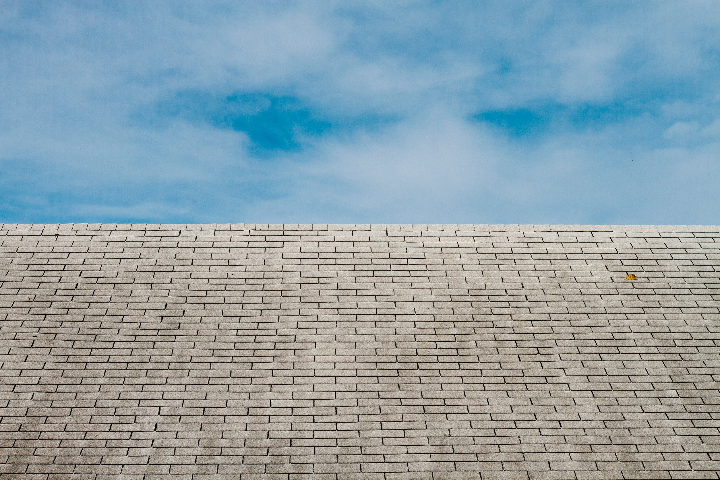 A sloped, light gray brick roof extends across the image under a bright blue sky with scattered clouds. A single yellow leaf rests on the roof near the upper right side.