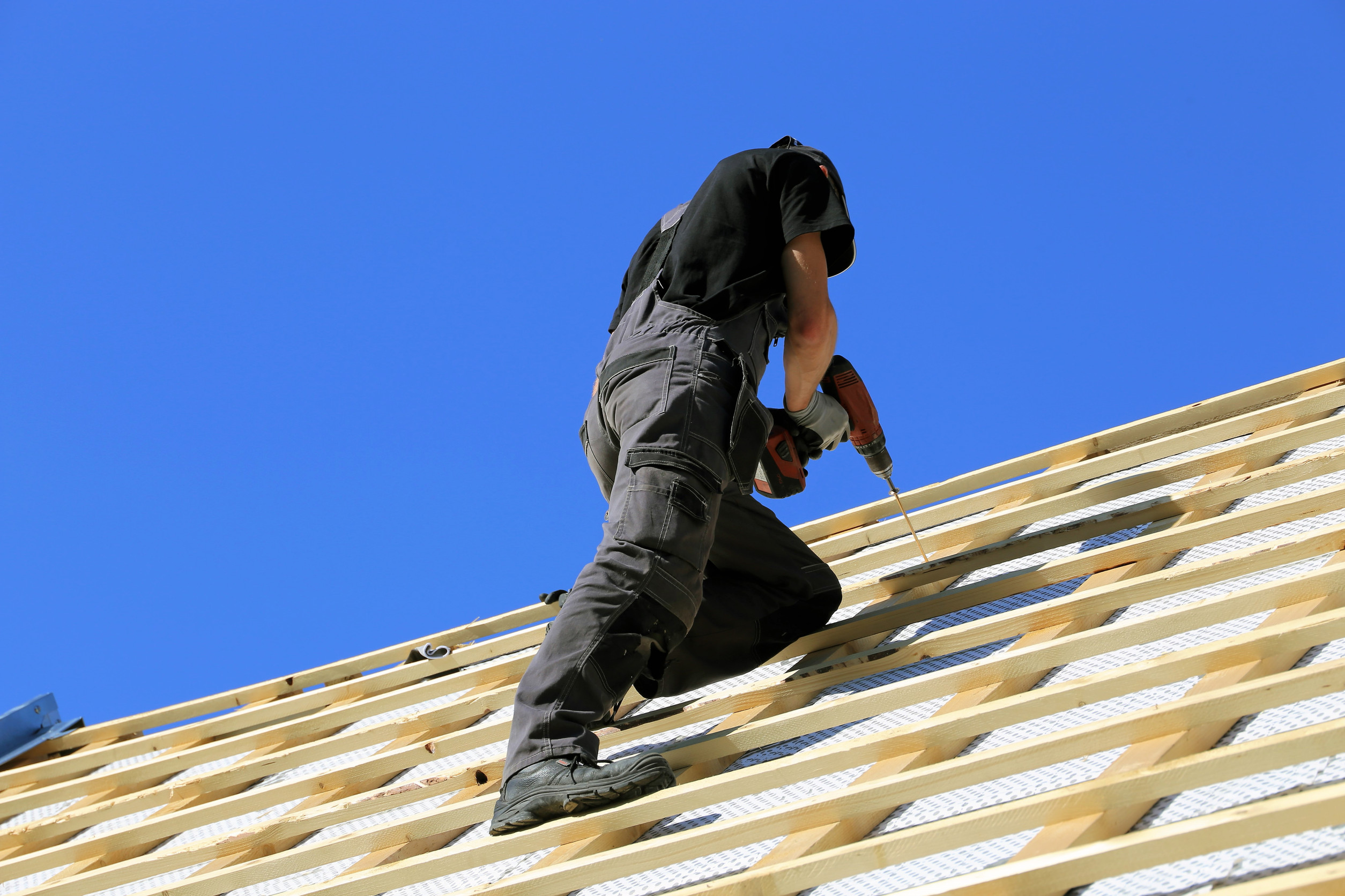 A person in work clothes and boots is using a power drill while installing materials on a sloped roof under a clear blue sky.