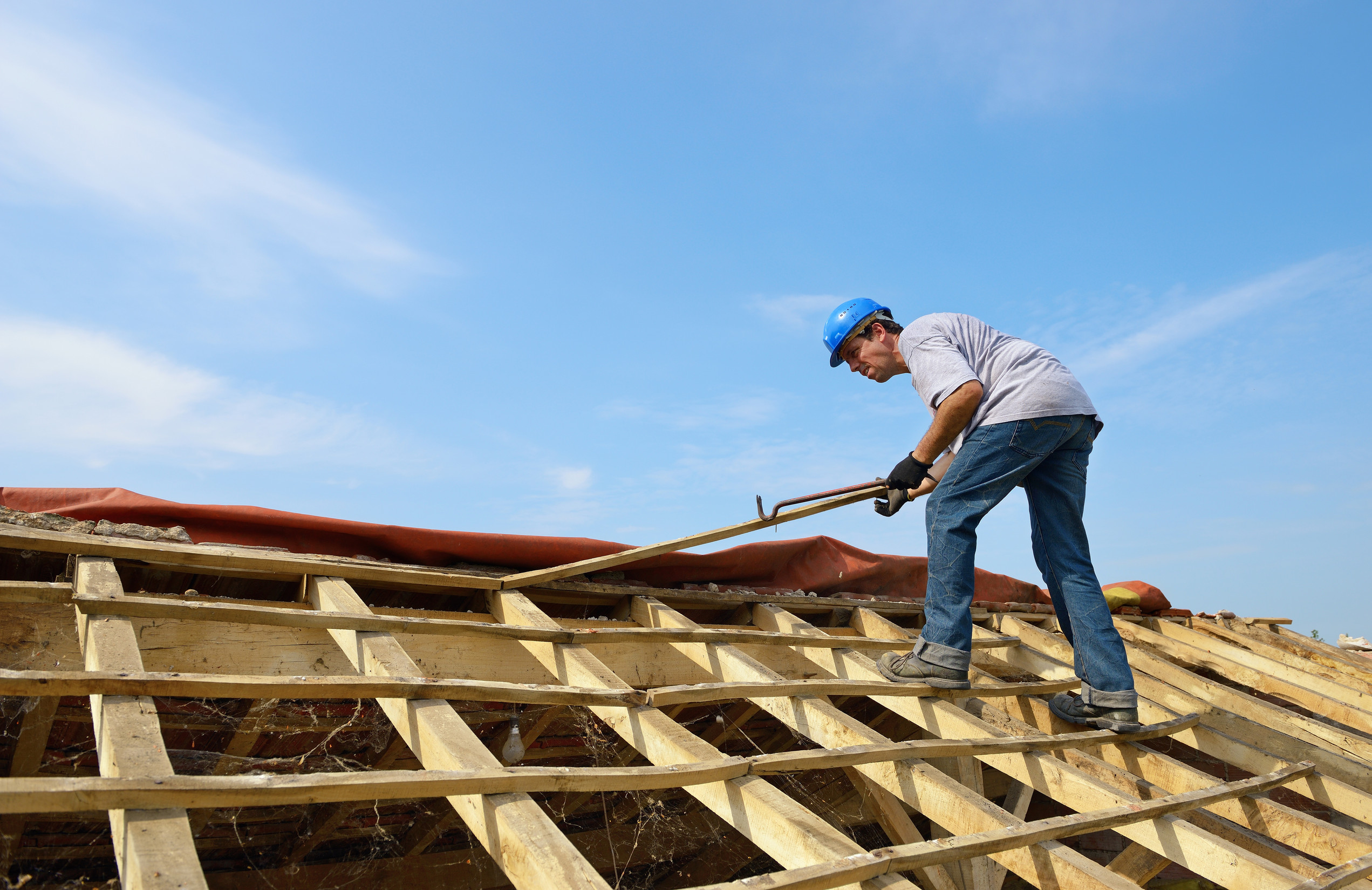 A construction worker in a blue hard hat and gloves uses a tool to work on the wooden framework of a roof under a clear blue sky.