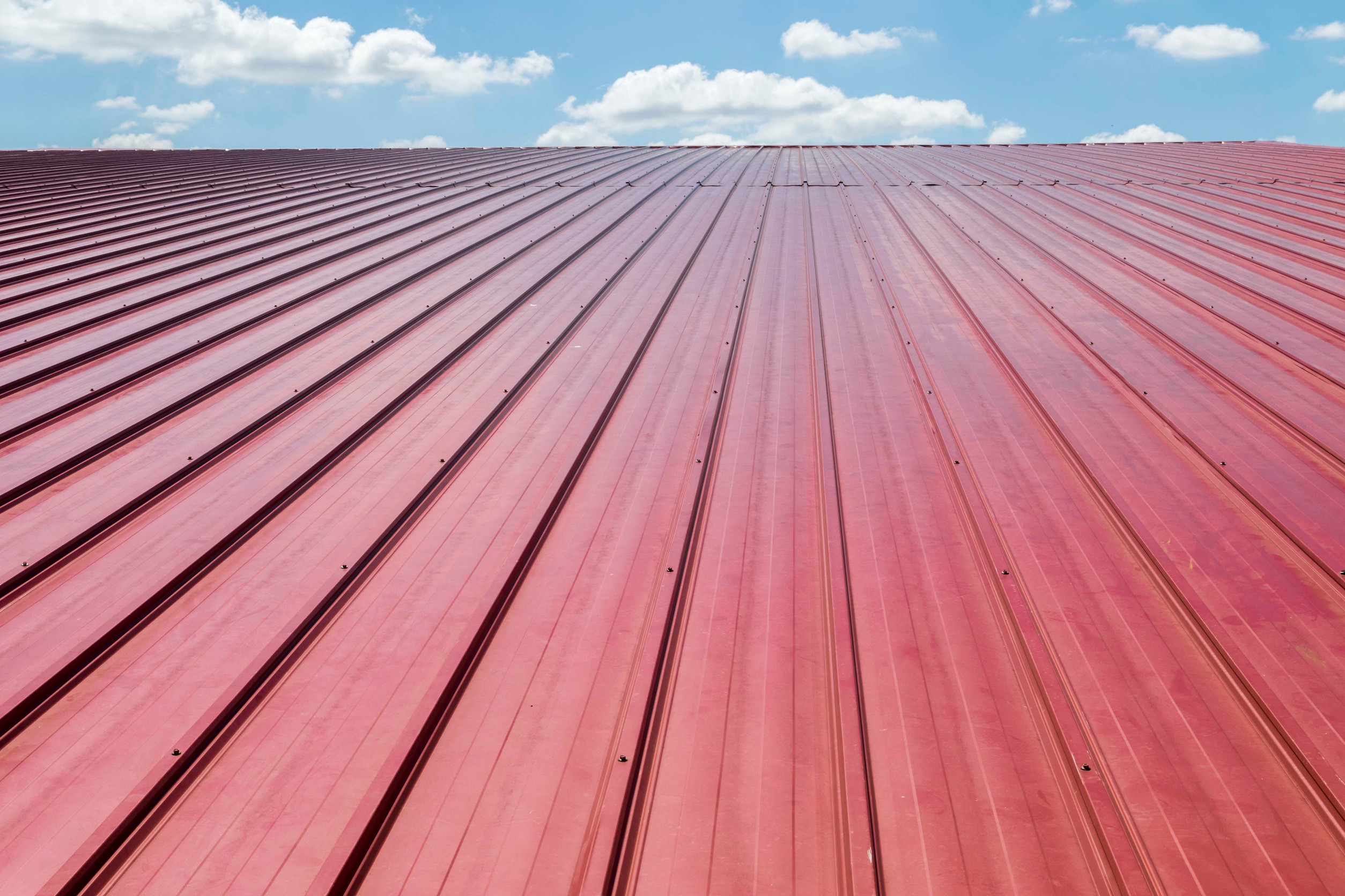 A close-up view of a red metal roof with ridged panels, photographed under a bright blue sky with scattered white clouds.