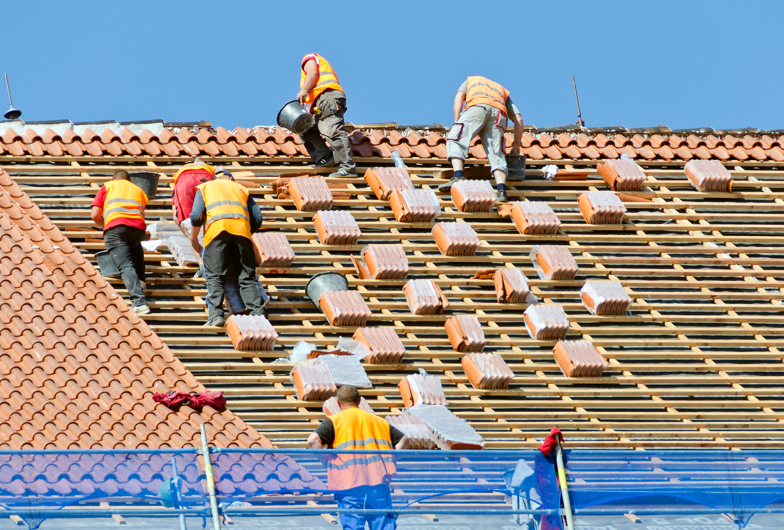 Several construction workers in orange safety vests are installing red roof tiles on a sloped roof under a clear blue sky. Stacks of tiles are distributed across the wooden beams of the roof.