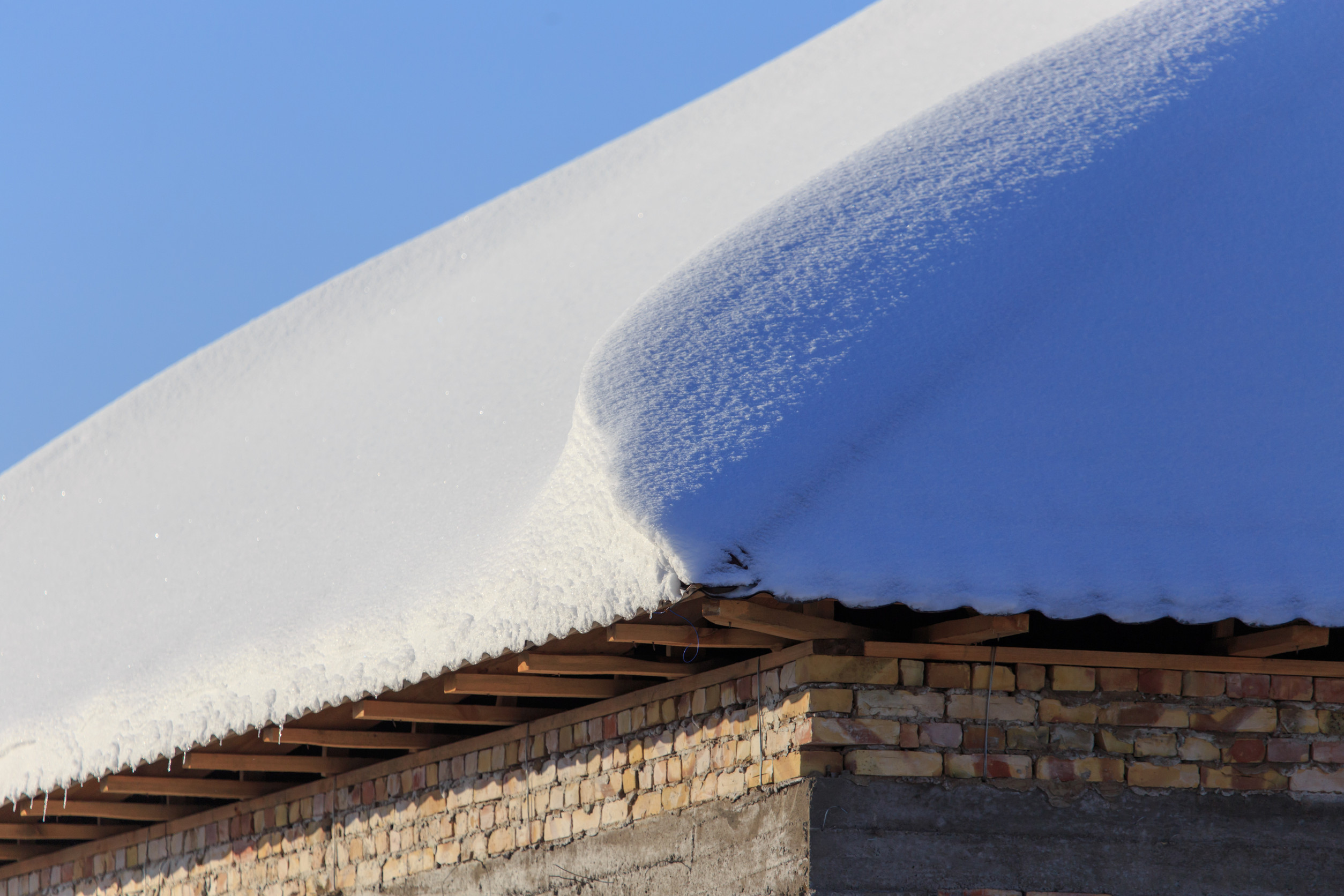 A thick layer of snow covers the sloped roof of a brick building under a clear blue sky. The snow has formed a smooth, curved edge along the roofline.