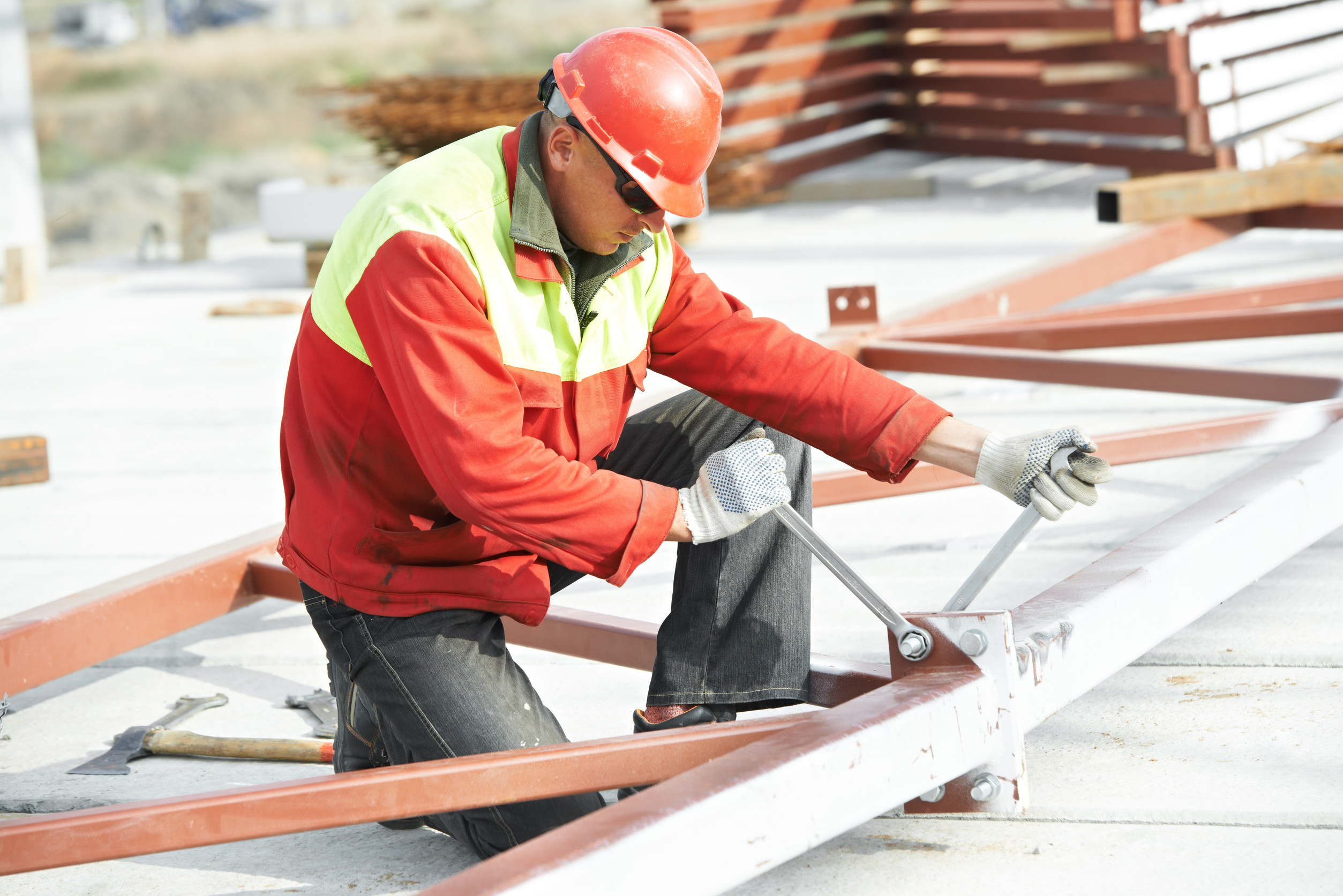 A construction worker wearing a red helmet and jacket tightens bolts on a steel frame with a wrench at a building site.