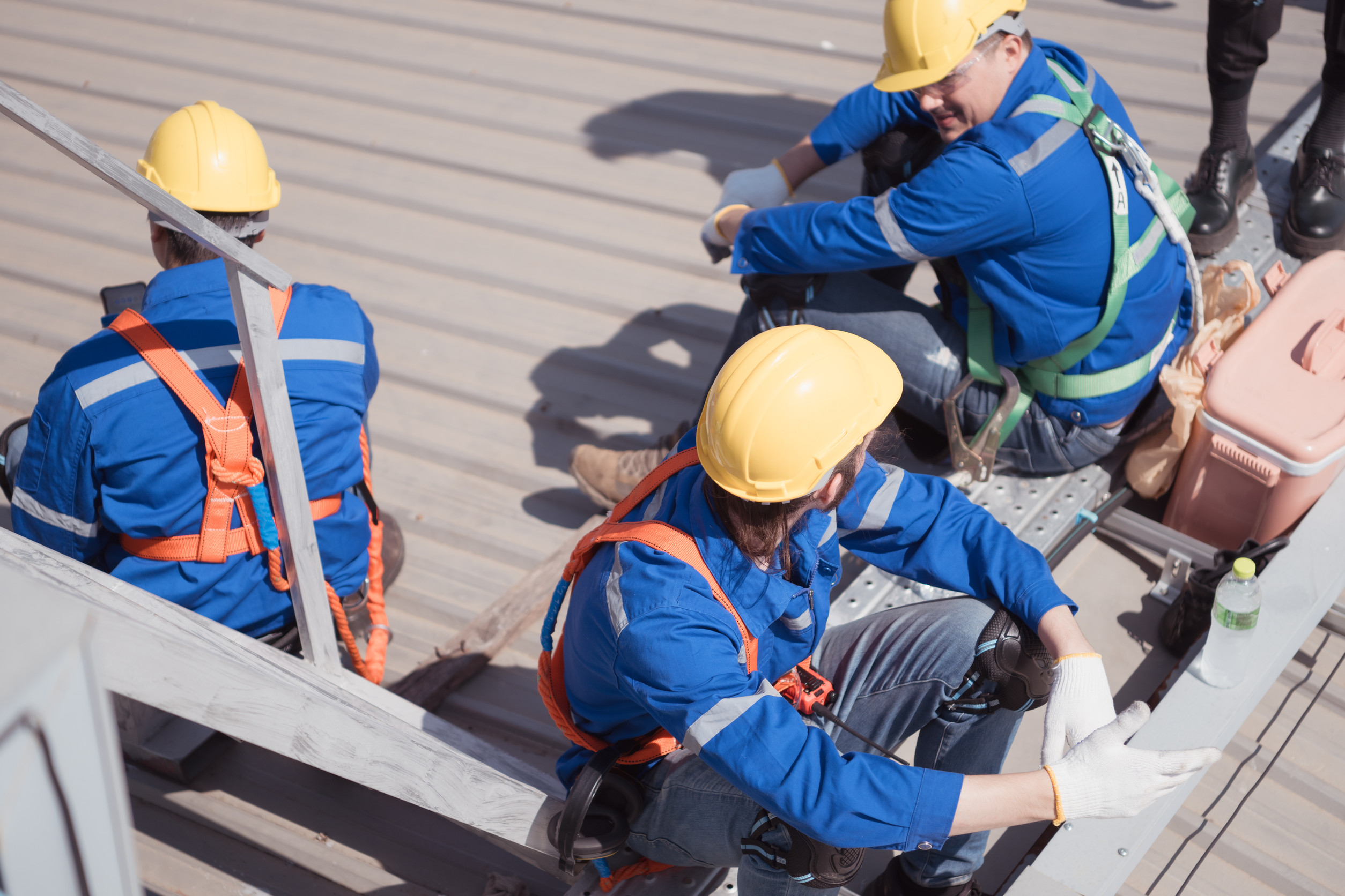 Three construction workers wearing yellow hard hats, blue uniforms, and safety harnesses sit and rest on a metal platform outdoors in the sunlight. Nearby are a pink toolbox and a bottle of drink.
