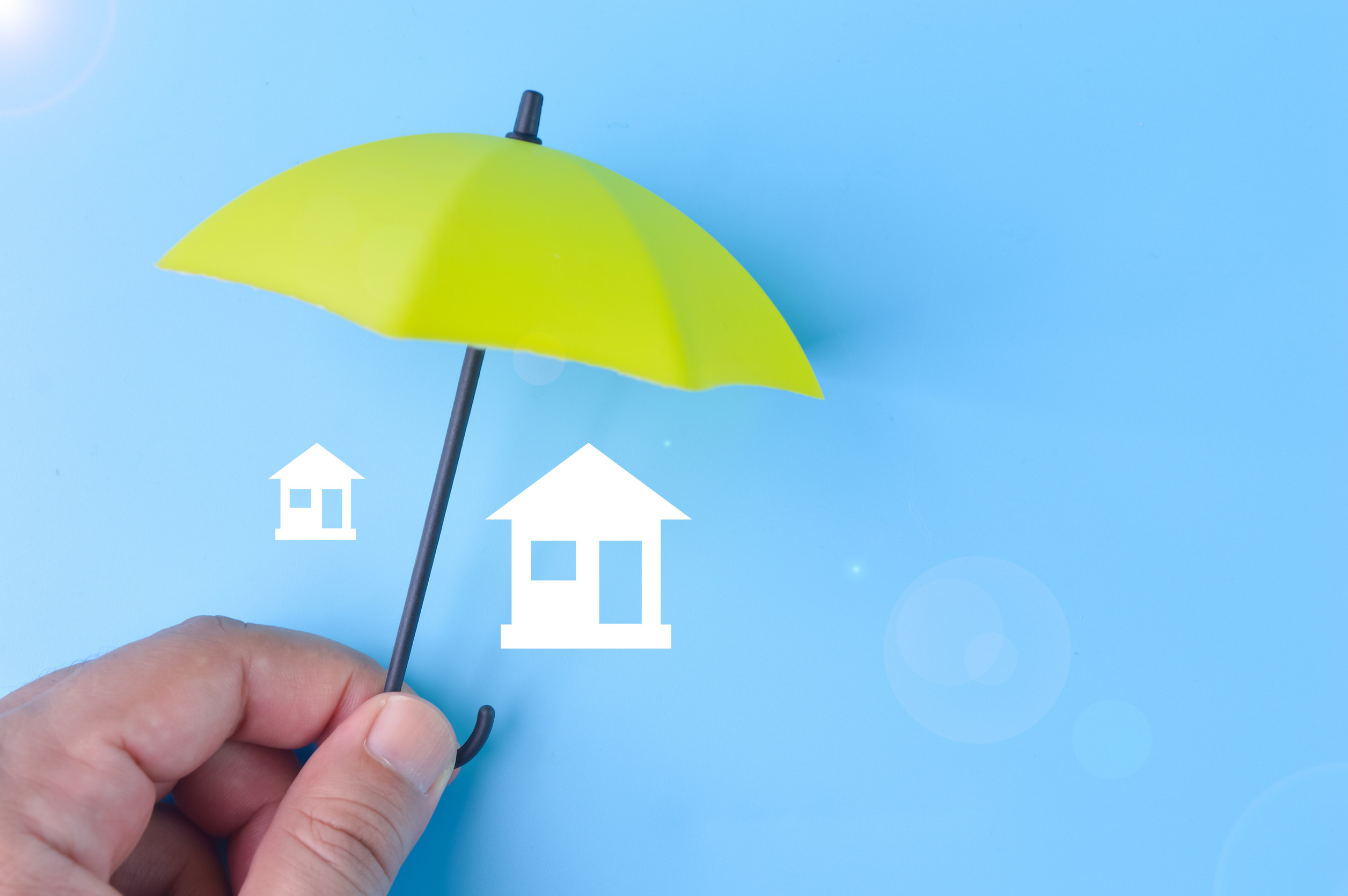 A hand holds a yellow umbrella over two white house icons on a blue background, symbolizing protection or home insurance.