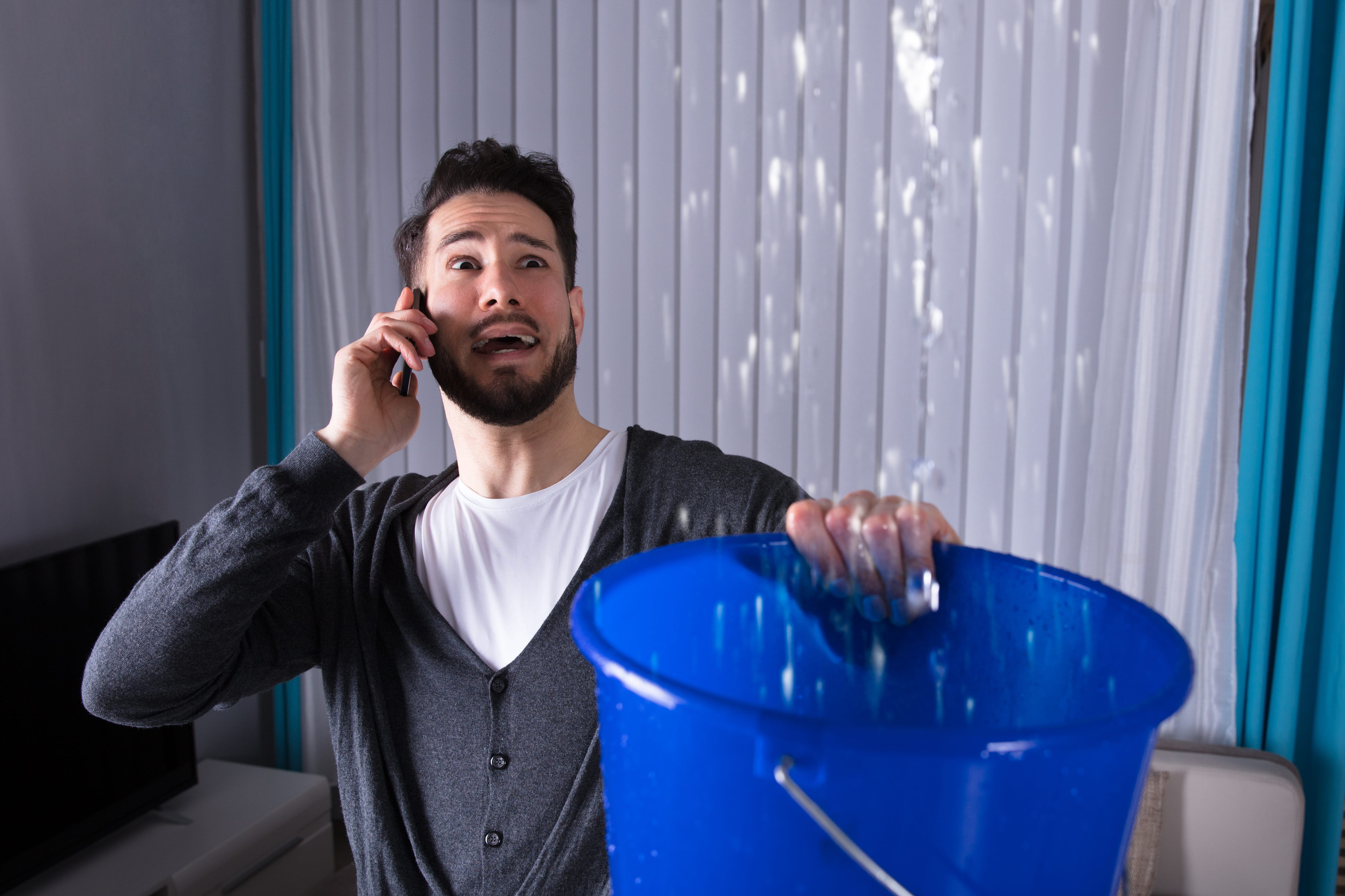 A worried man holding a blue bucket under a ceiling leak talks on the phone, likely seeking help for the water problem in his home.